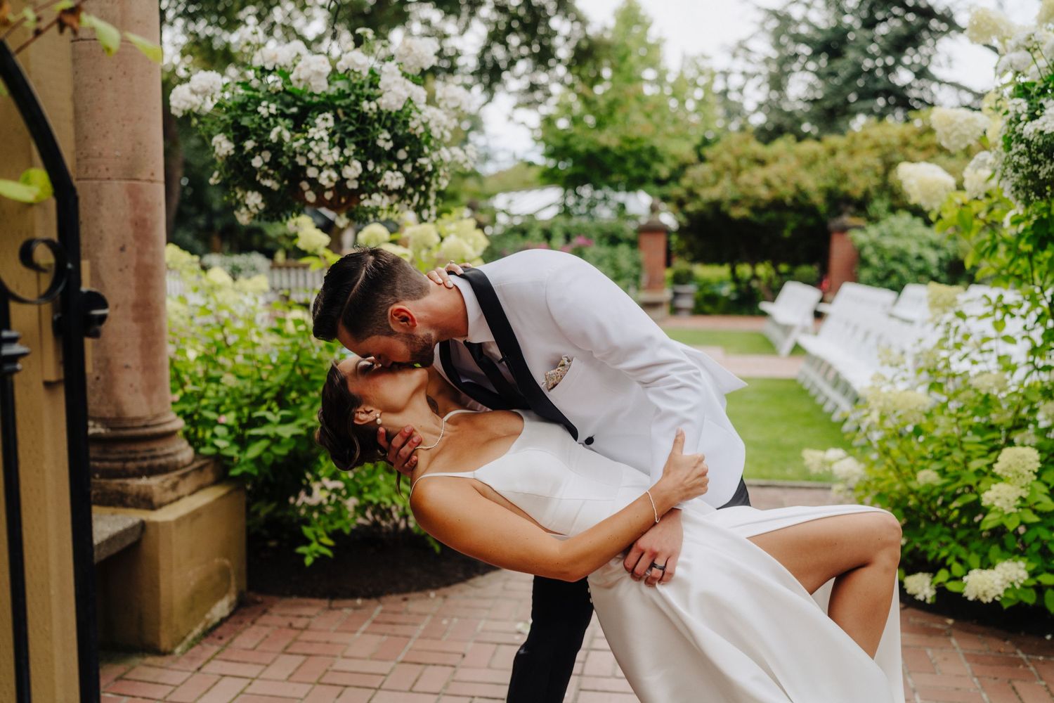 A romantic outdoor garden wedding ceremony with white chairs and floral arrangements along the aisle.