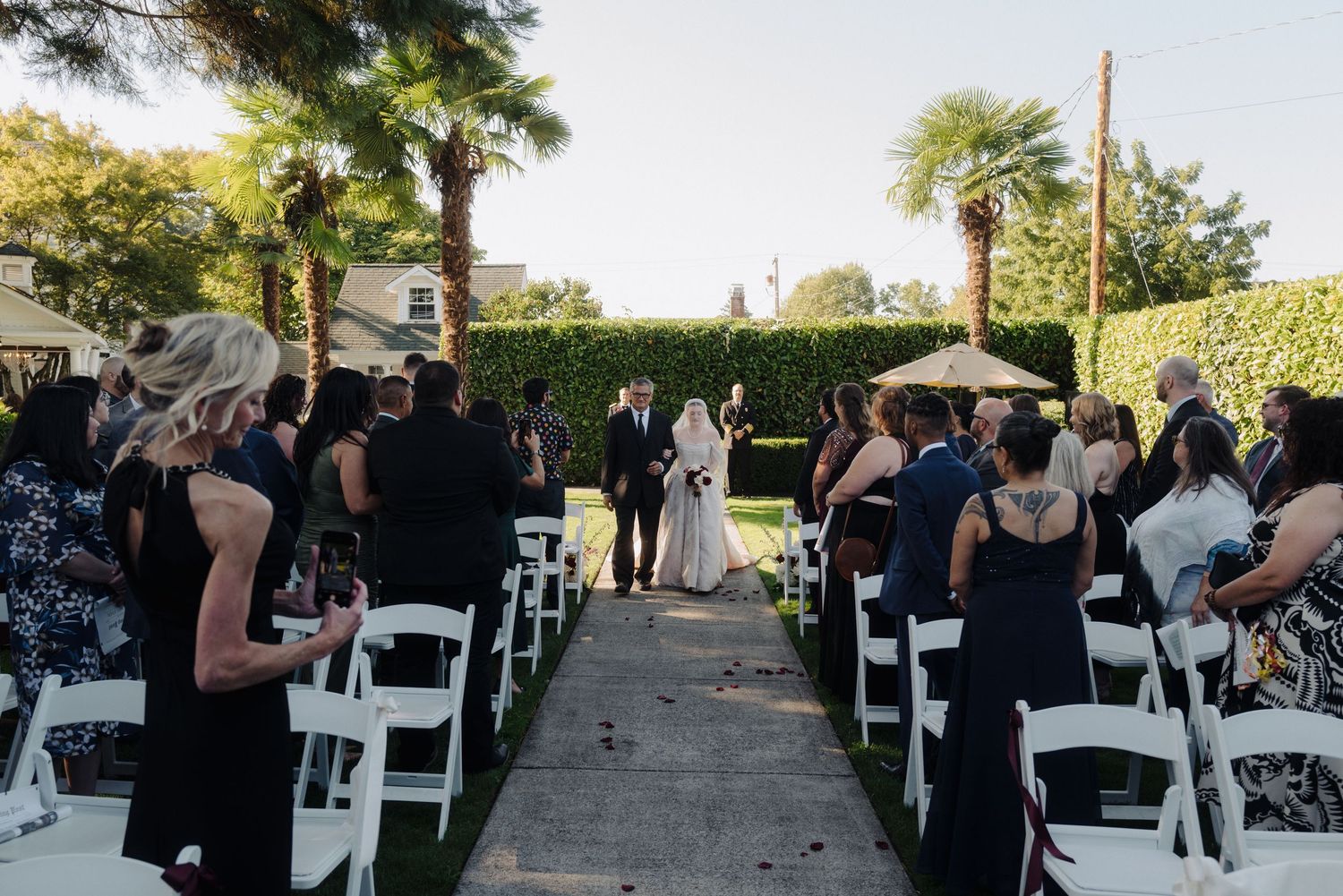 Wedding guests in dark attire seated in white chairs during an outdoor ceremony.