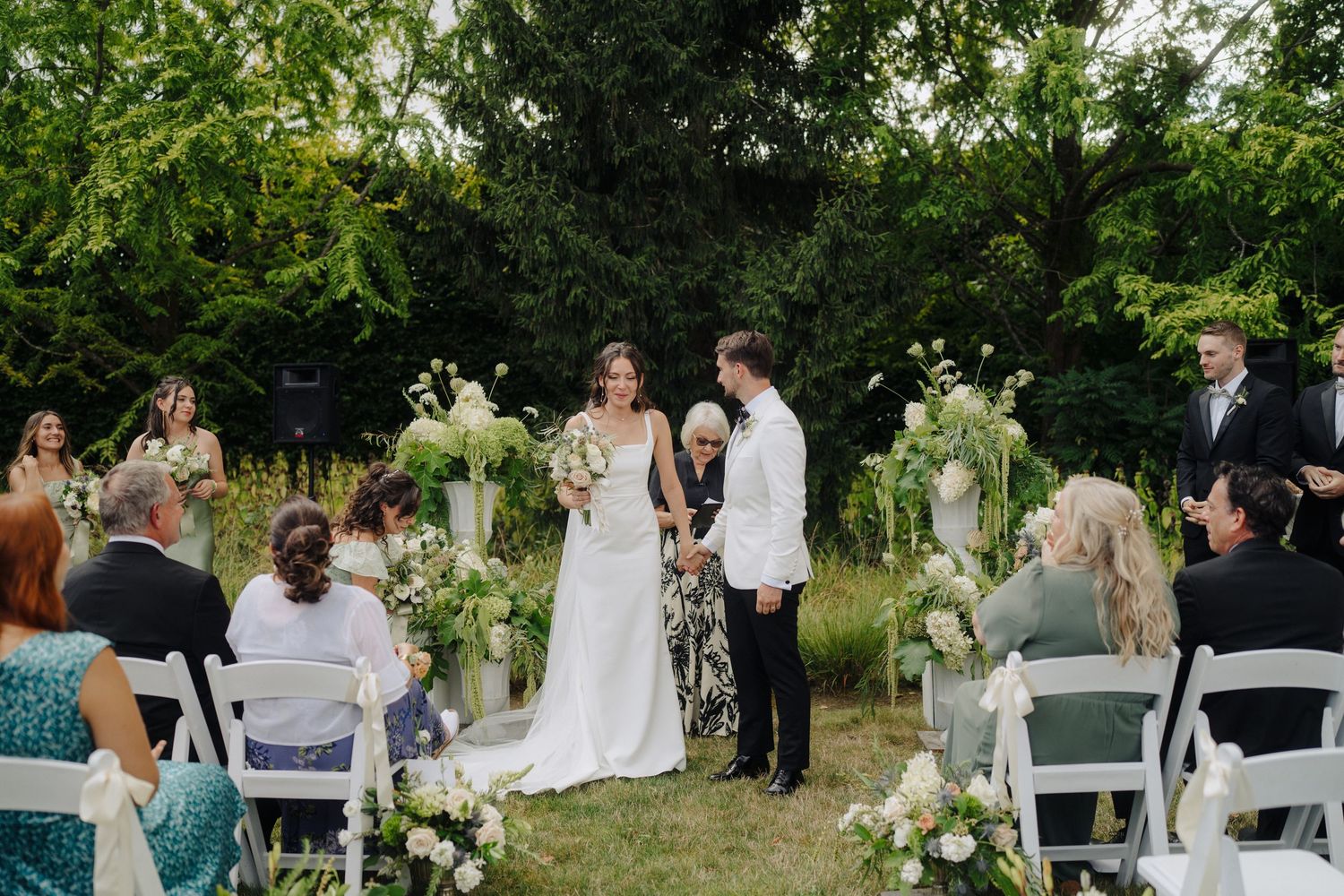 Wedding ceremony guests gathered outdoors with white chairs and floral decorations.
