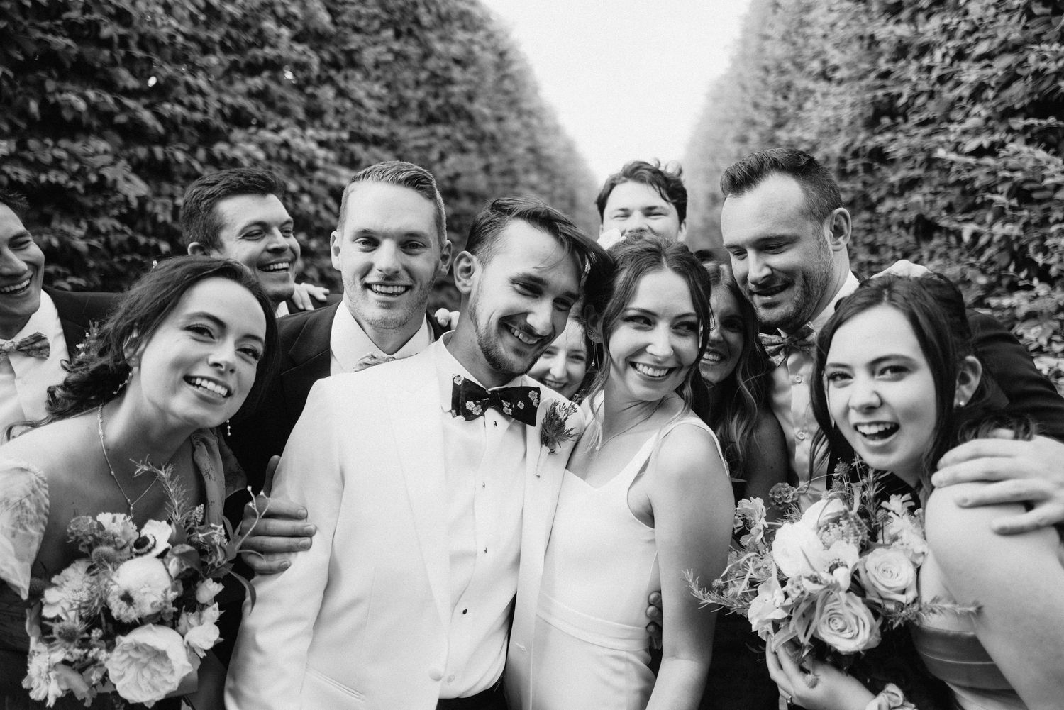 Candid group photo of wedding party members laughing together outdoors with greenery in background.