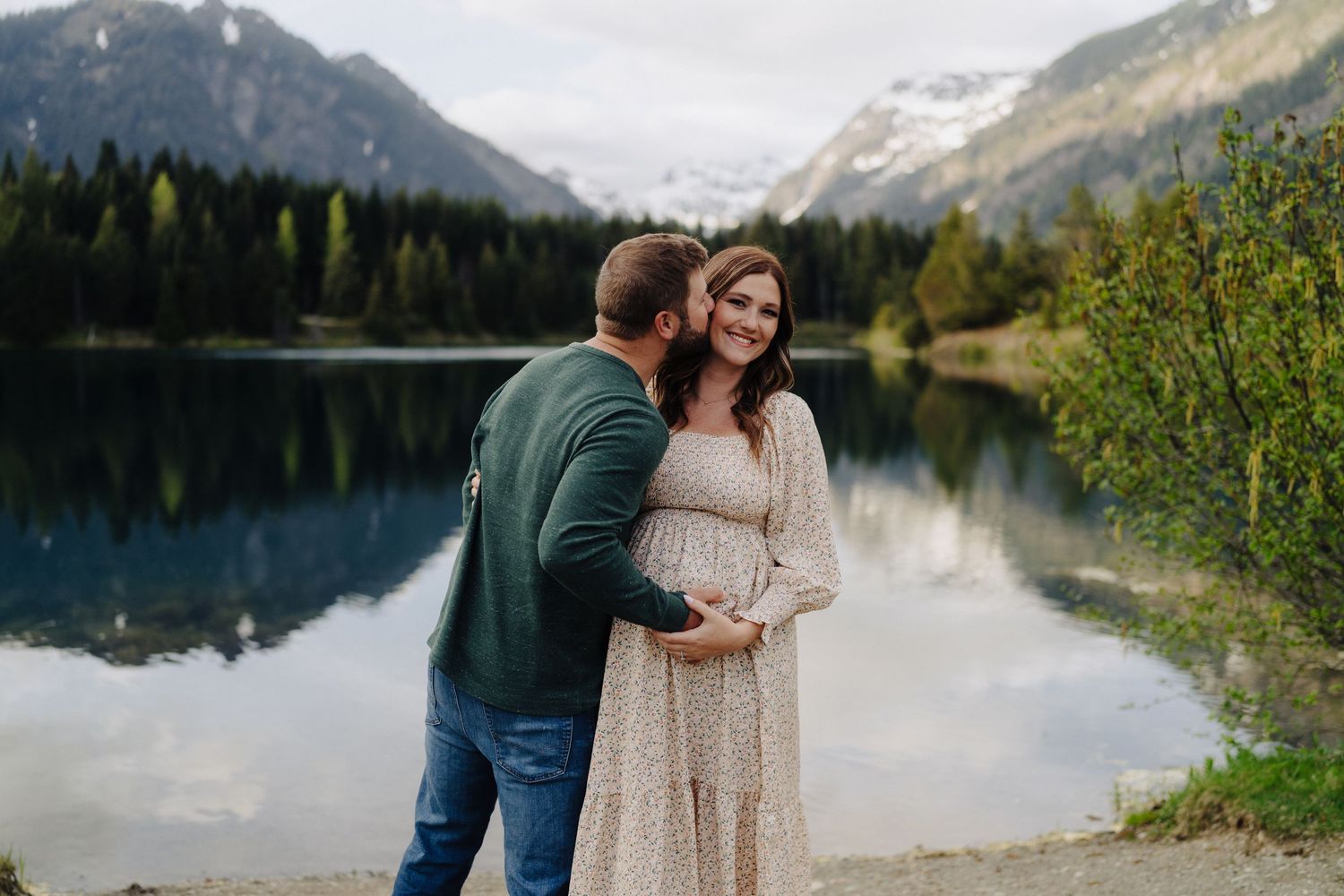 A couple stands on a rocky shore with snow-capped mountains and a serene lake in the background.