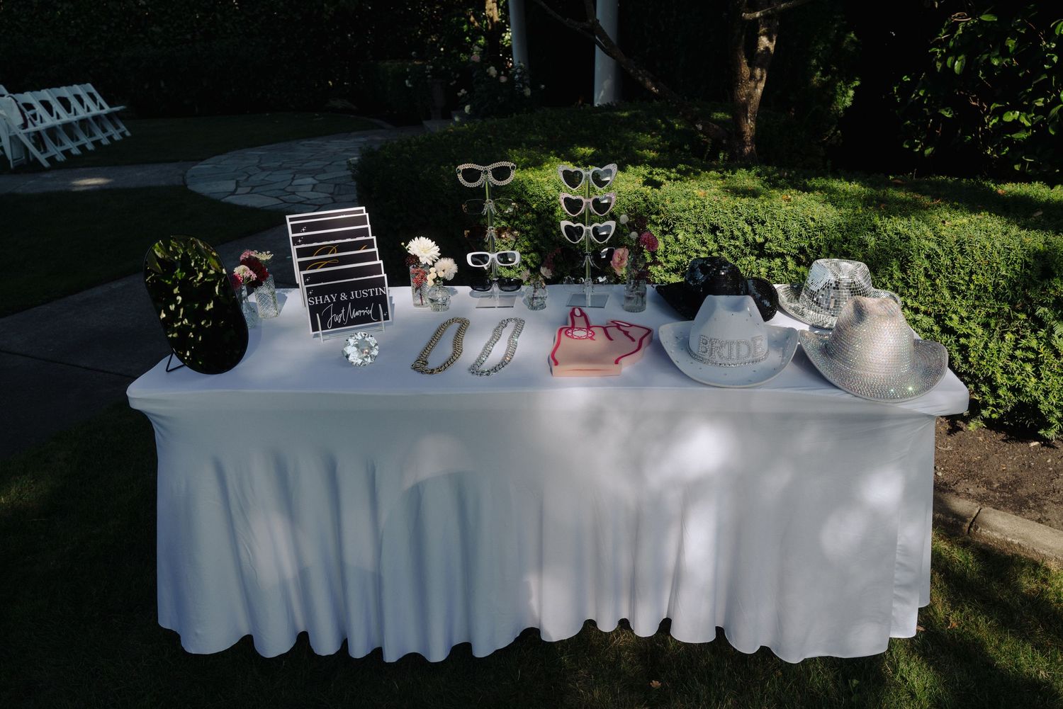 A white-clothed reception table displays wedding decor and refreshments in an outdoor garden setting.