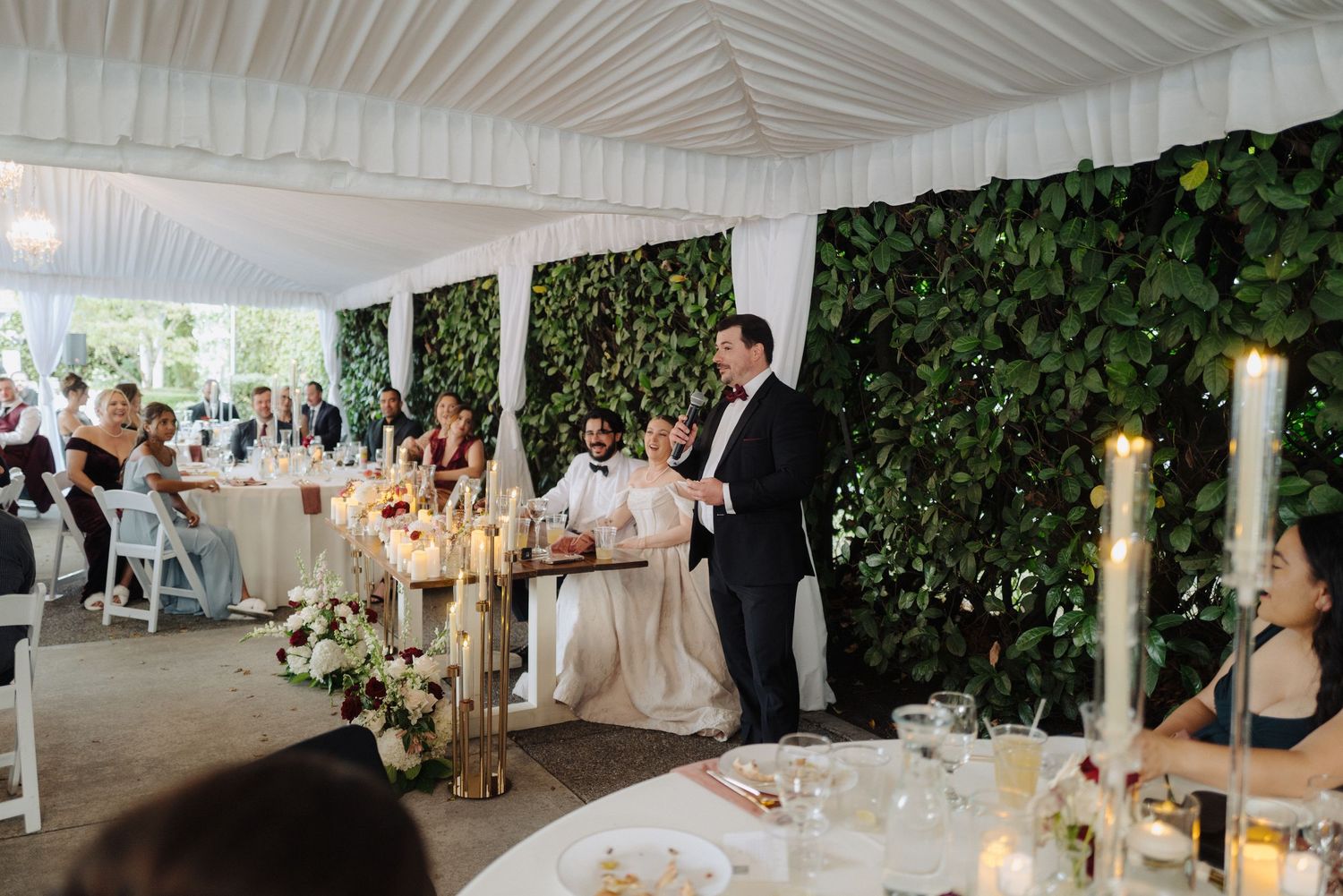 An elegant white wedding tent reception space decorated with greenery walls and crystal chandeliers.
