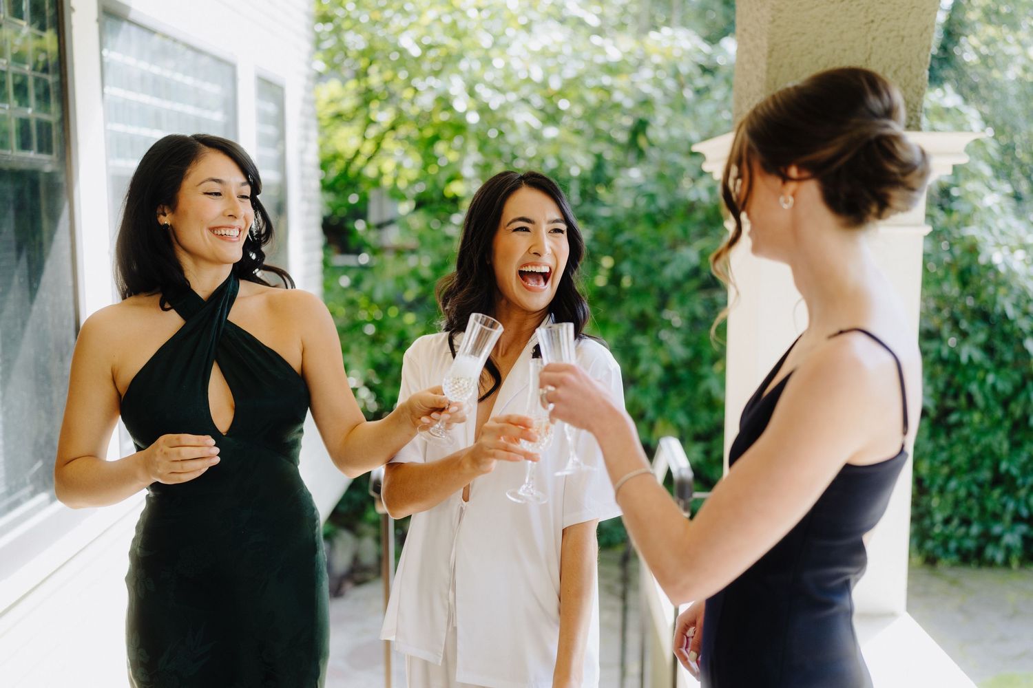 Three friends in elegant dresses share drinks and laughter at an outdoor gathering.