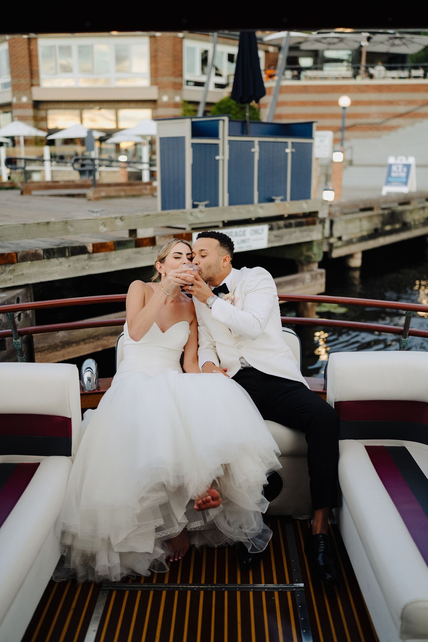 Bride and groom sipping champagne together while seated on a luxury boat docked at a waterfront venue.