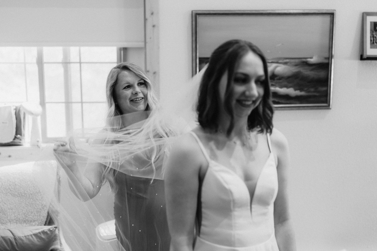 A candid black and white moment showing a bride in a white dress while someone adjusts her veil.