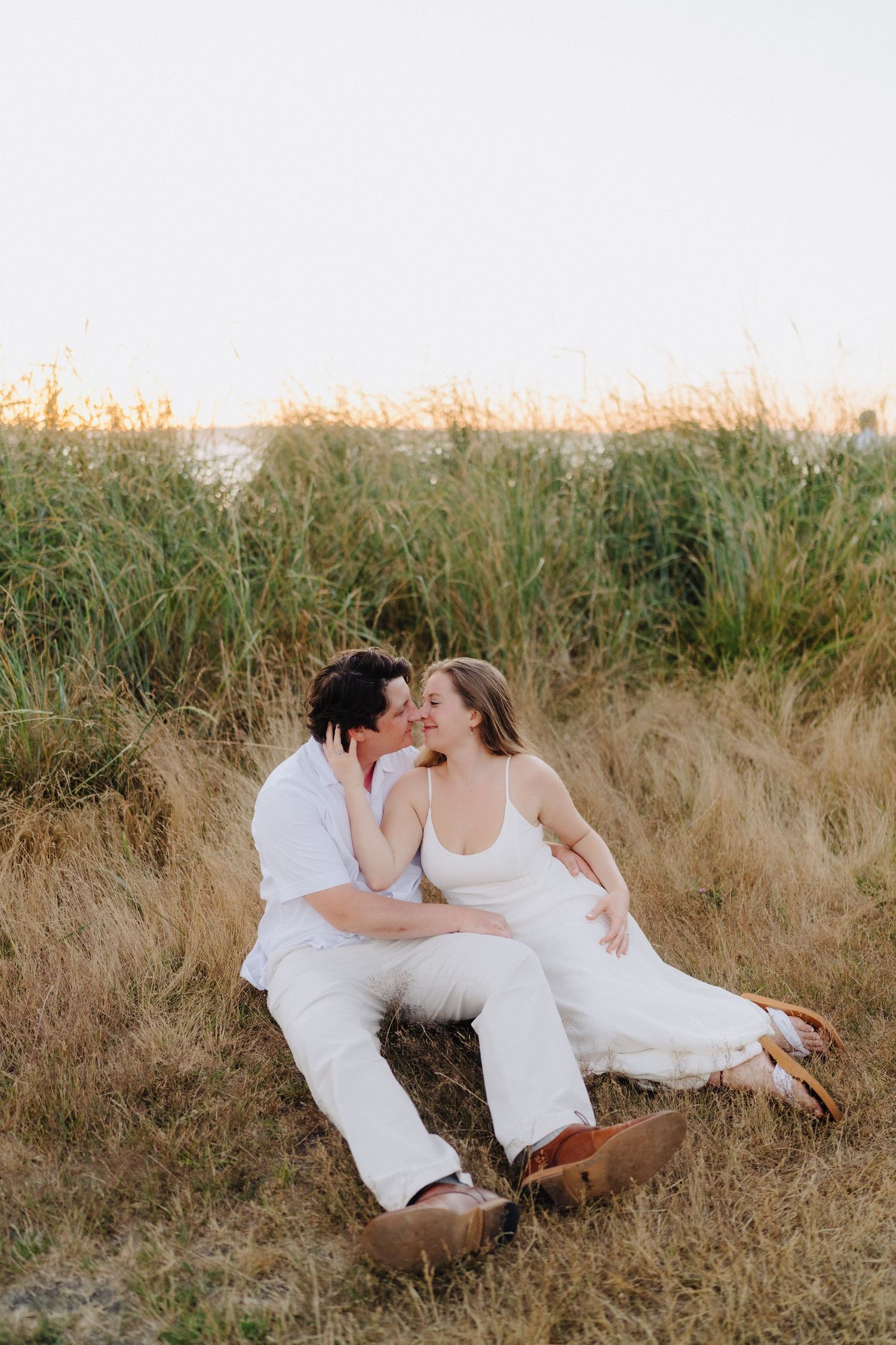 A couple in white outfits shares tender moments while sitting in tall grass during sunset.