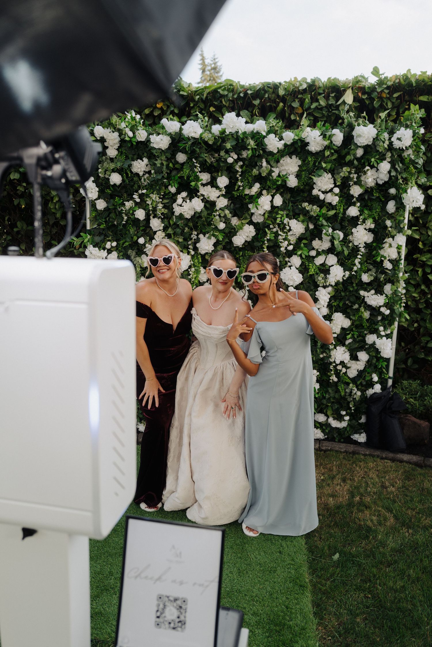 Three women posing in front of a floral wall backdrop at a wedding.