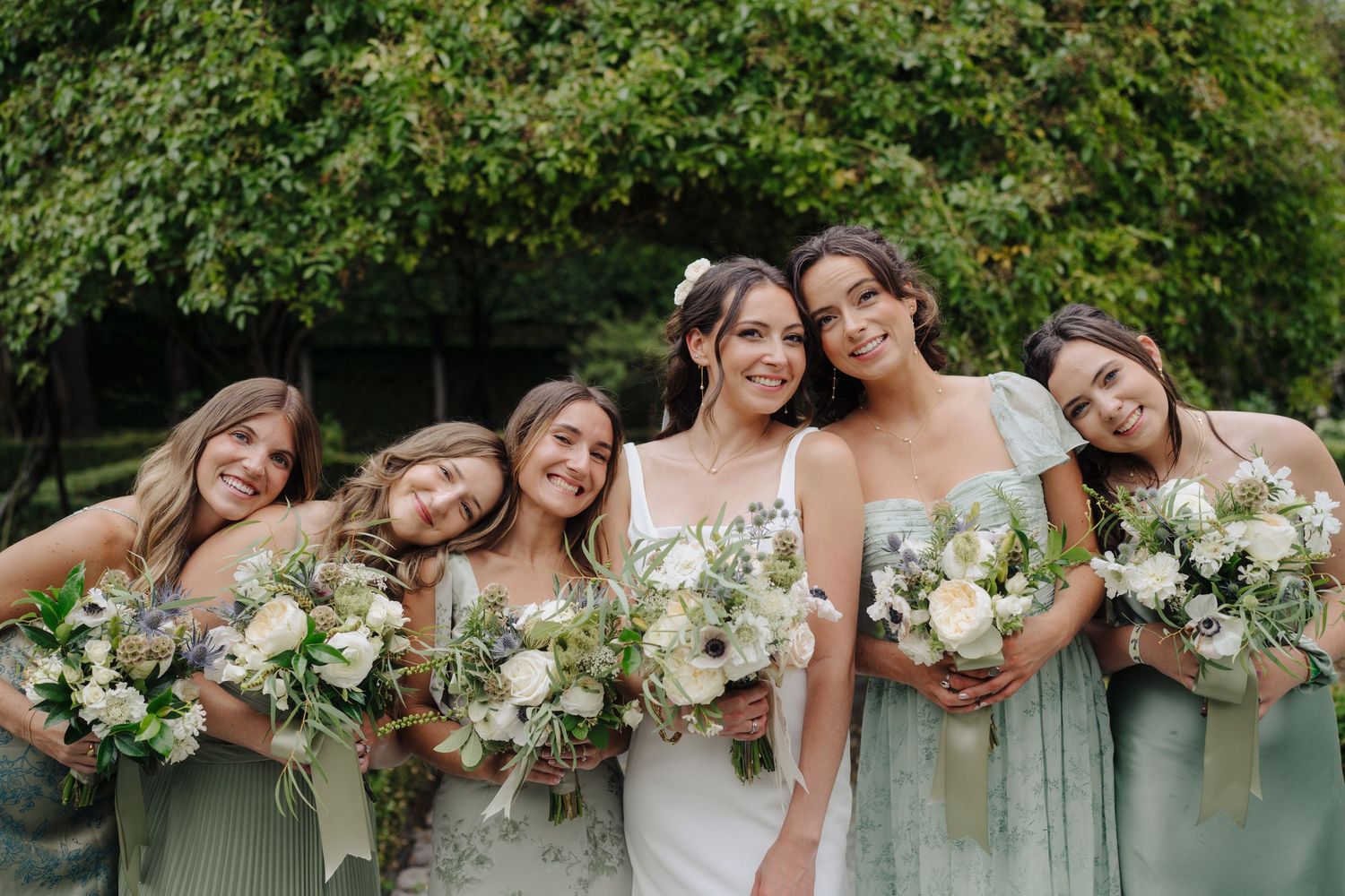 Bridesmaids in matching dresses pose with bouquets against a green hedge.