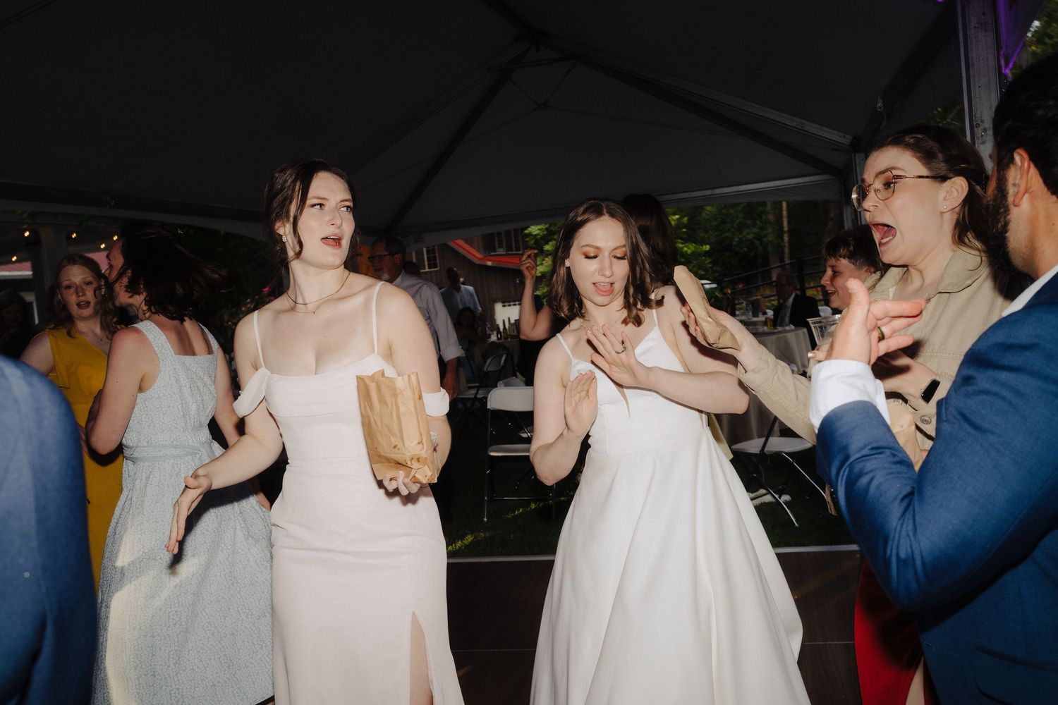 Two brides in white dresses dancing joyfully with guests at a wedding reception under a tent.