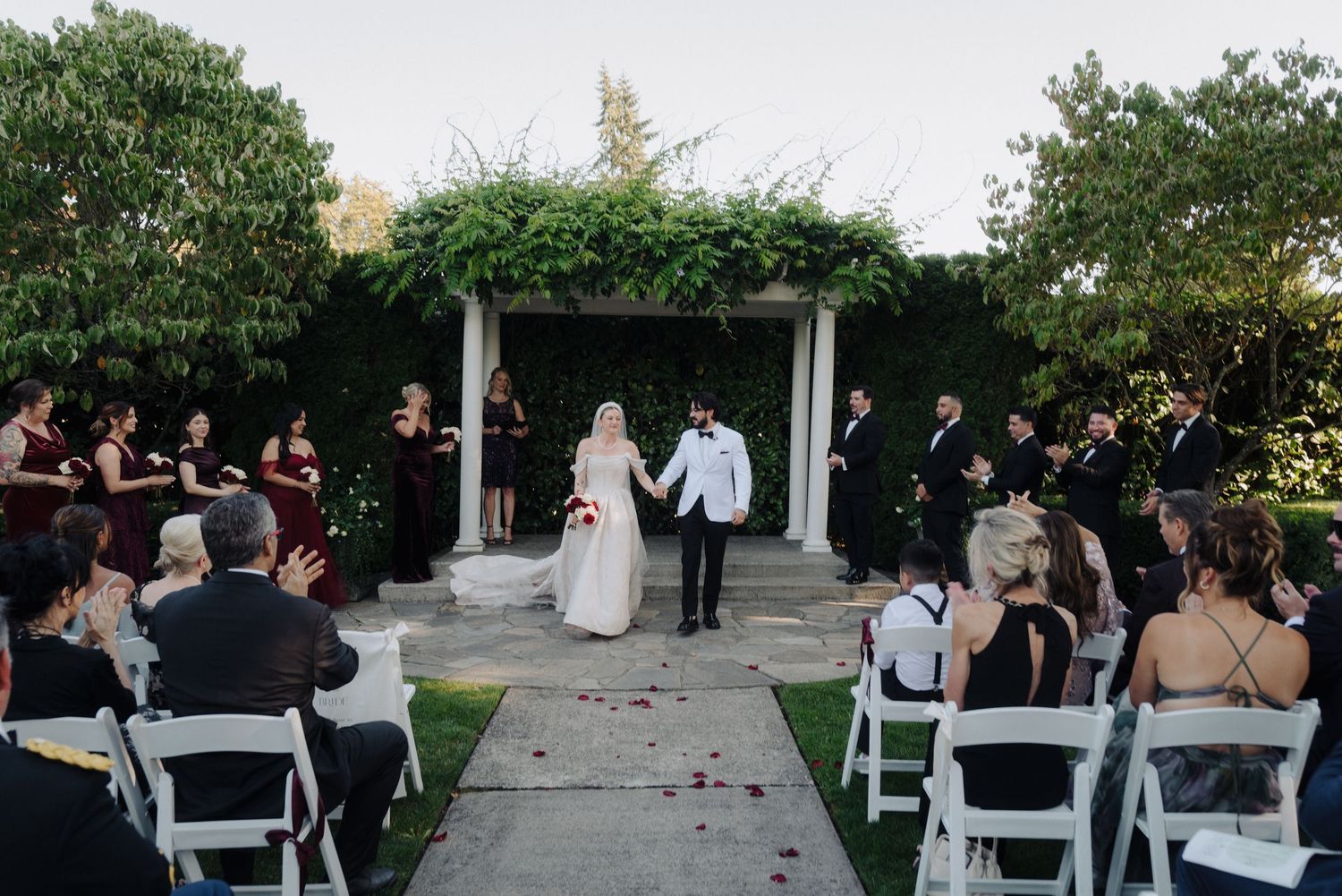 Wide angle shot of wedding ceremony with guests seated in white chairs surrounded by lush landscaping.