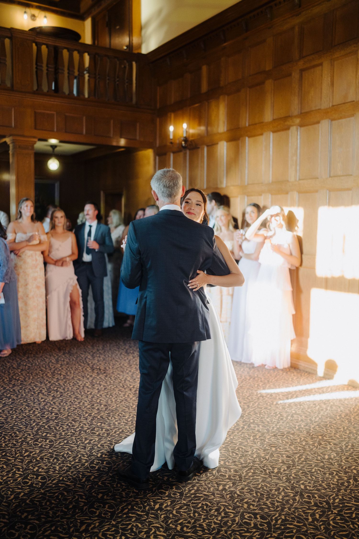Wedding guests dancing in an elegant wood-paneled ballroom with patterned carpet.