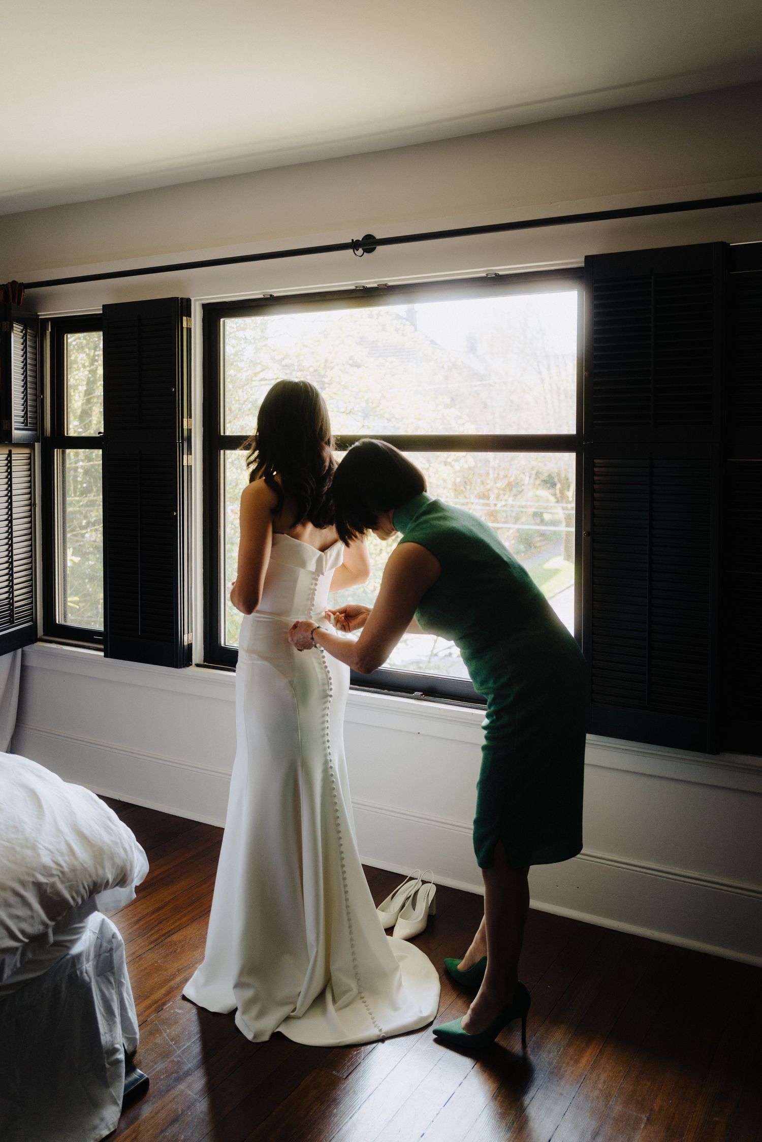 Two figures stand by a window in a bedroom as morning light streams in during wedding preparations.