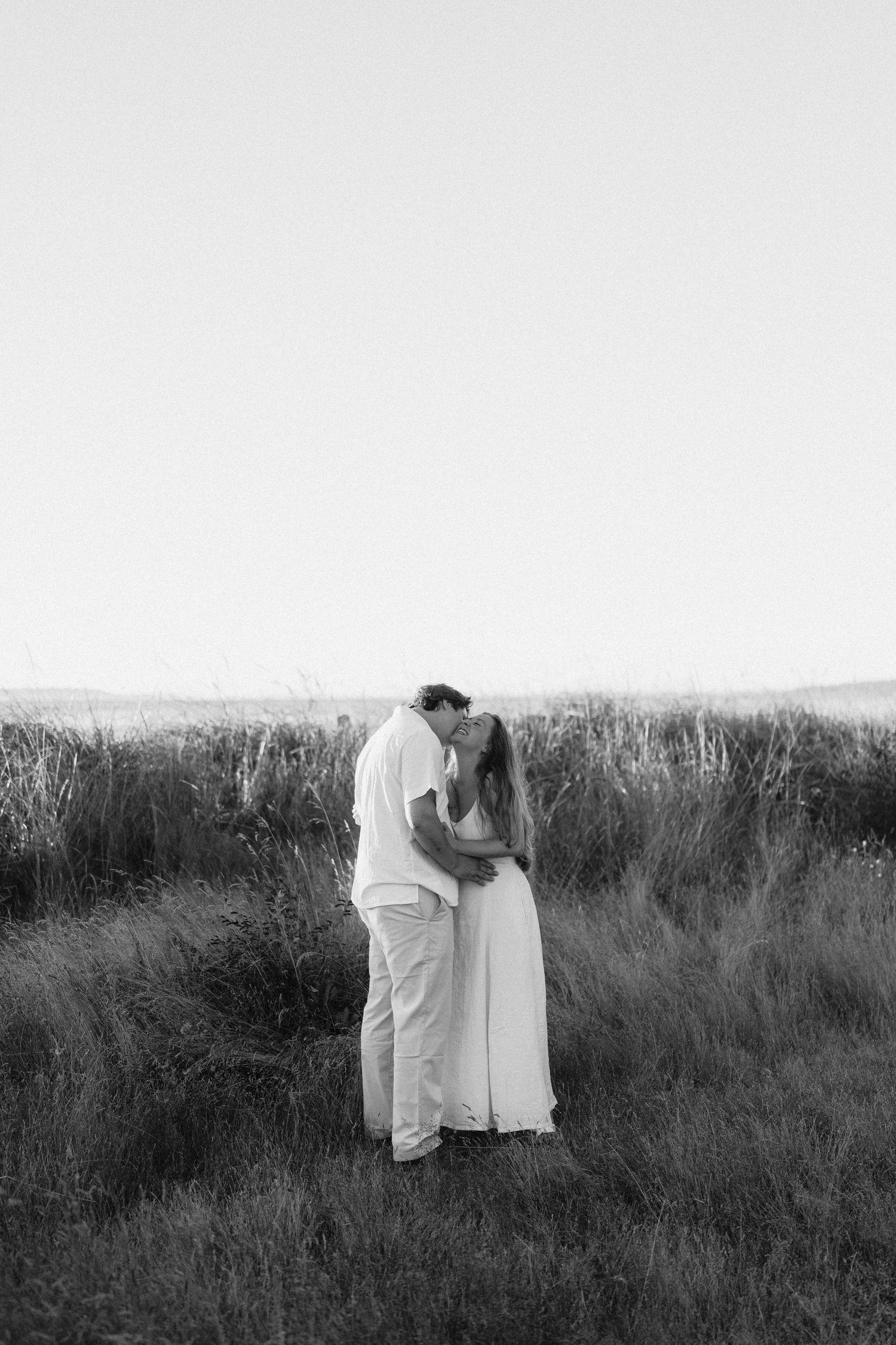 A romantic couple in white clothing embraces in a field of tall grass at sunset.