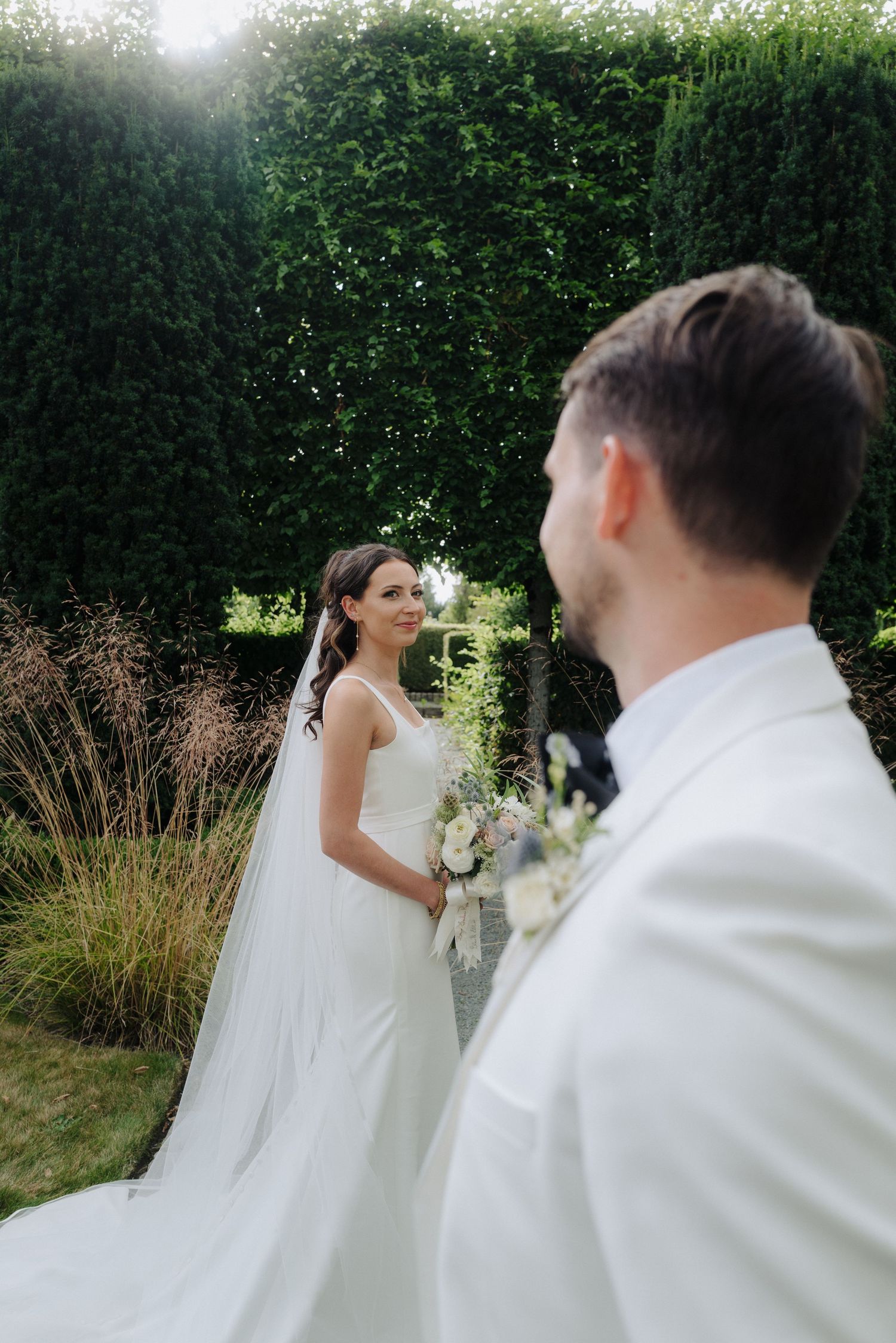 Wedding couple poses together in an outdoor garden setting with soft sunlight filtering through trees.