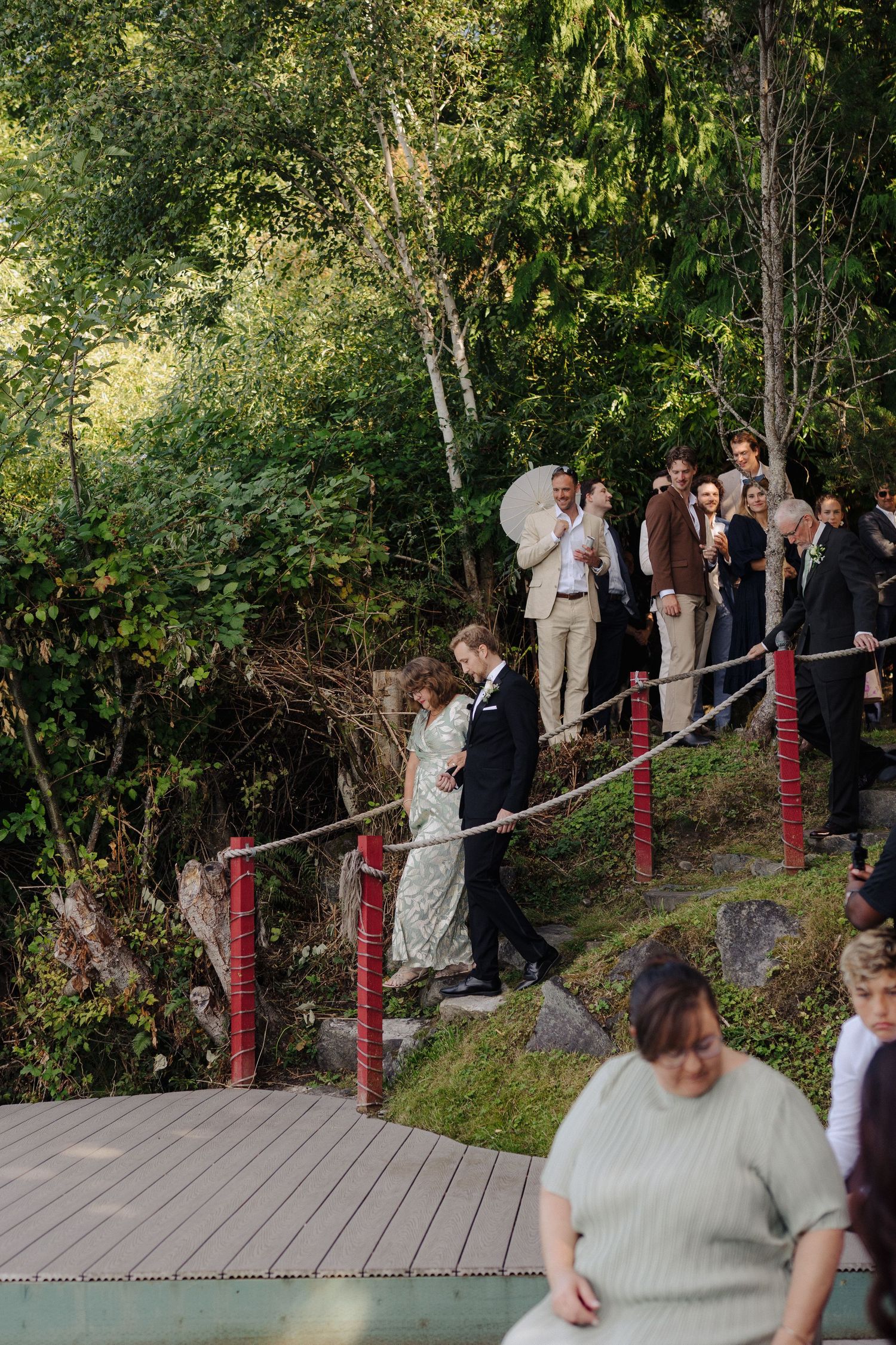Wedding guests gather on a wooden deck with red railings surrounded by lush greenery.