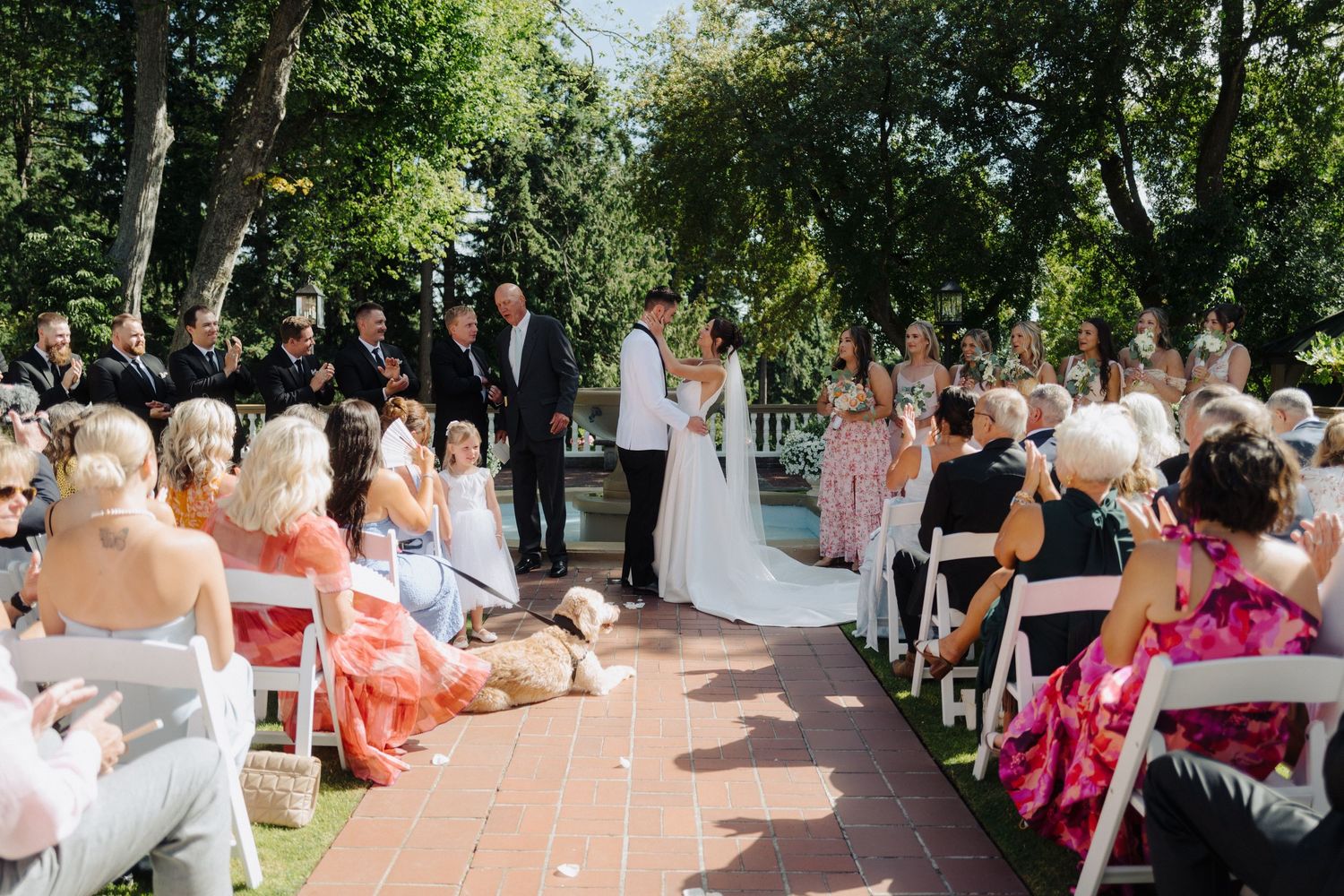 Outdoor wedding ceremony guests seated in rows of white chairs along a garden path surrounded by greenery.