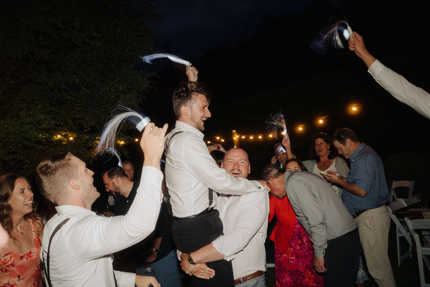 Wedding reception guests dance under evening sky with string lights.