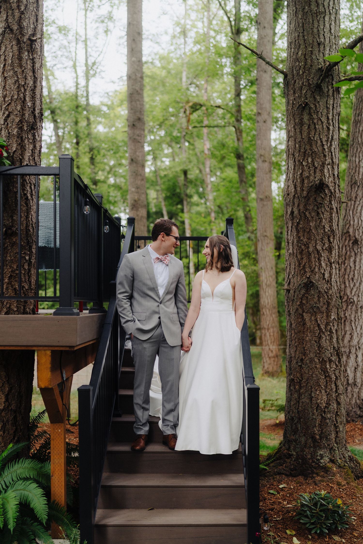 Couple descends wooden stairs surrounded by tall pine trees and ferns in a forest setting.