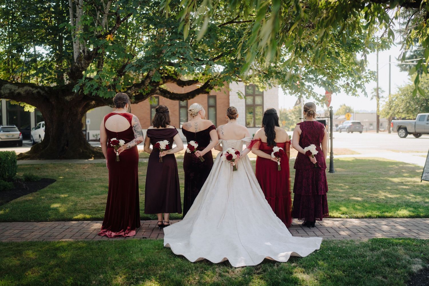 Wedding party in burgundy dresses and white gown pose together on a grassy lawn at an outdoor venue.
