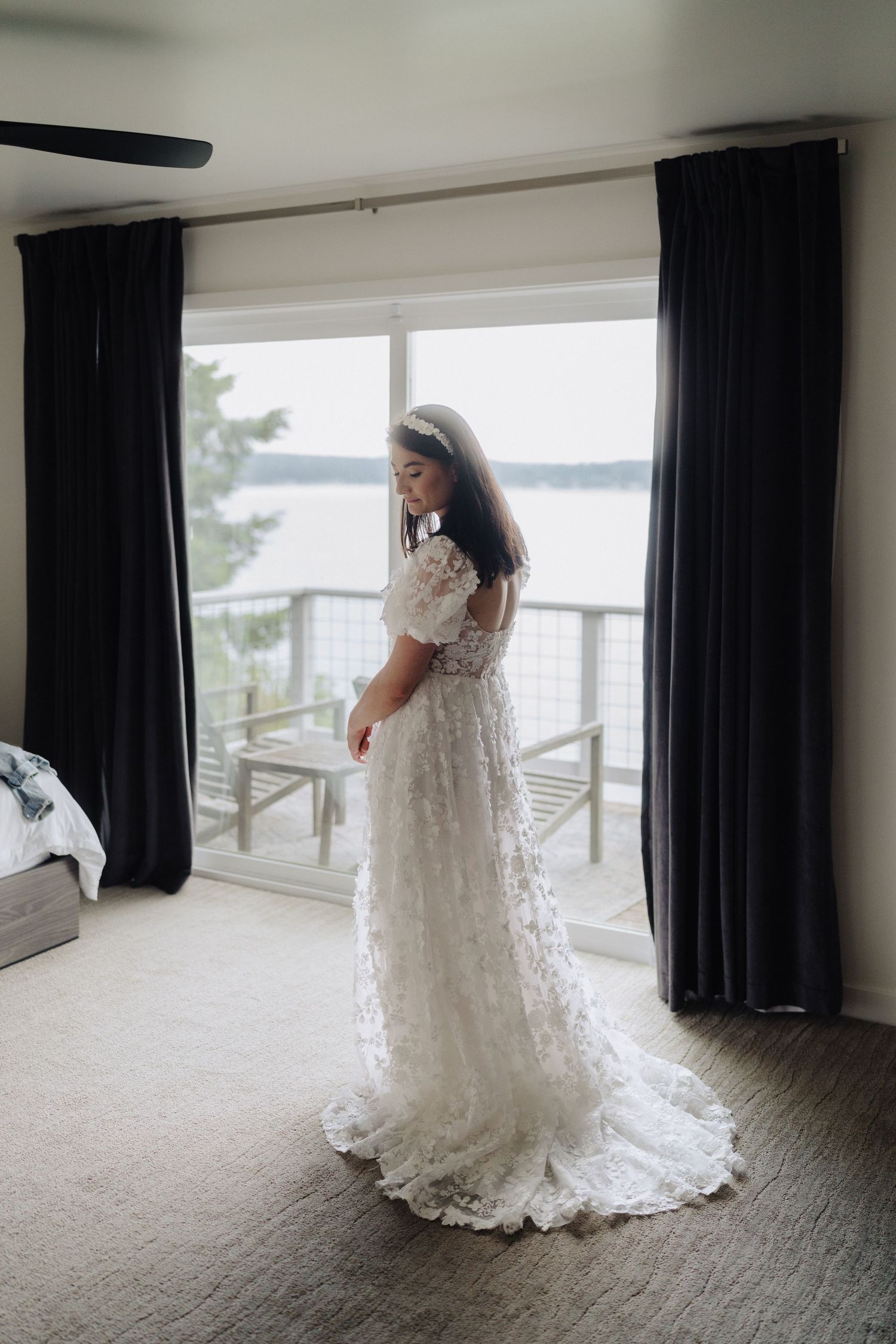 A lace wedding dress is showcased in a room with large windows overlooking a waterfront view.