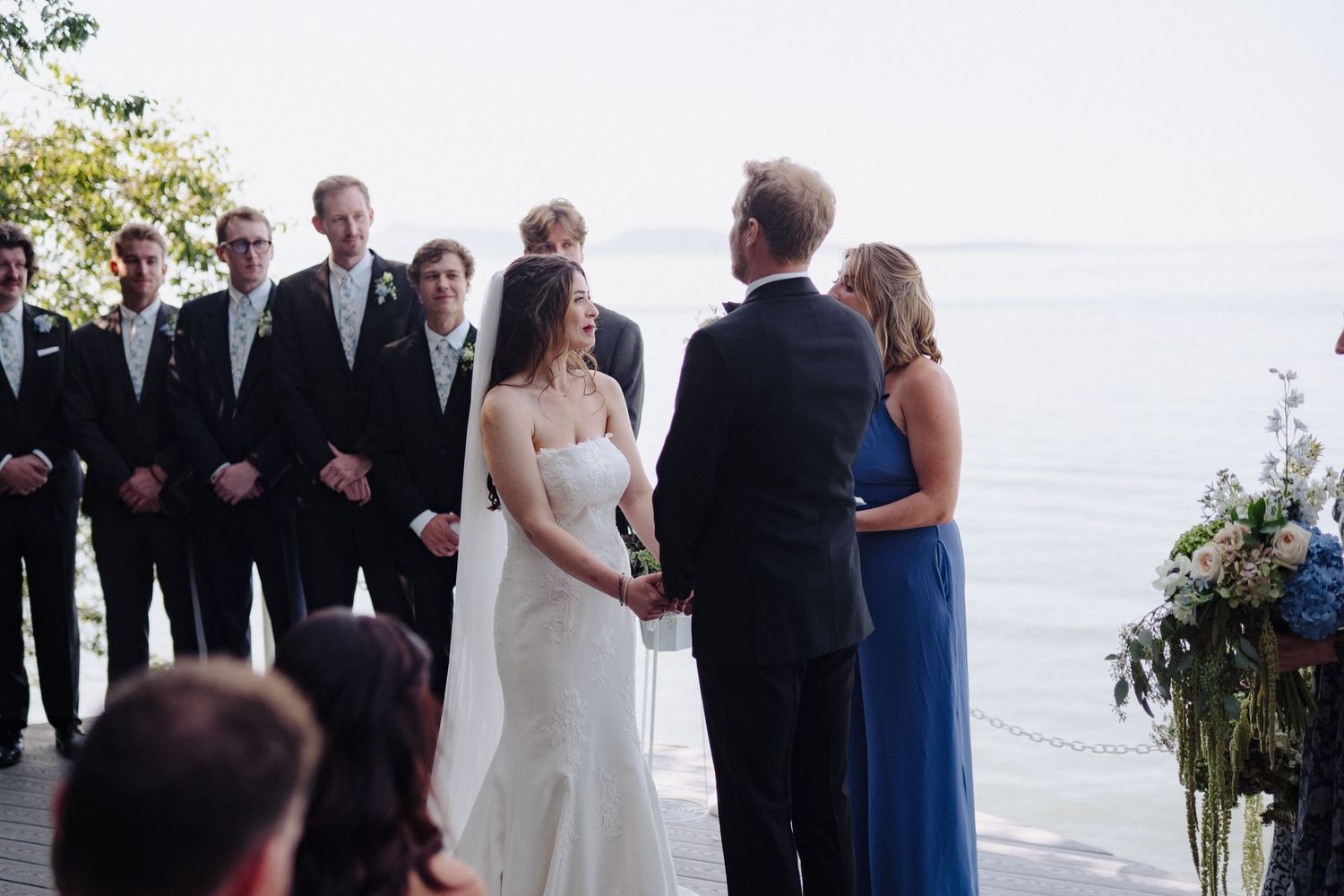 An outdoor wedding ceremony takes place with guests gathered by the ocean at sunset.