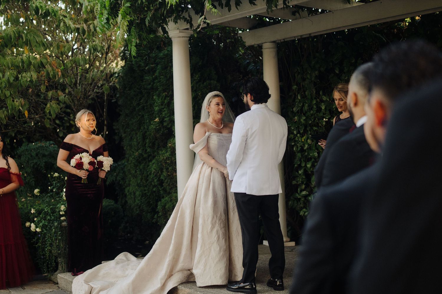 Wedding ceremony taking place in a lush garden setting with white columns and greenery decorations.
