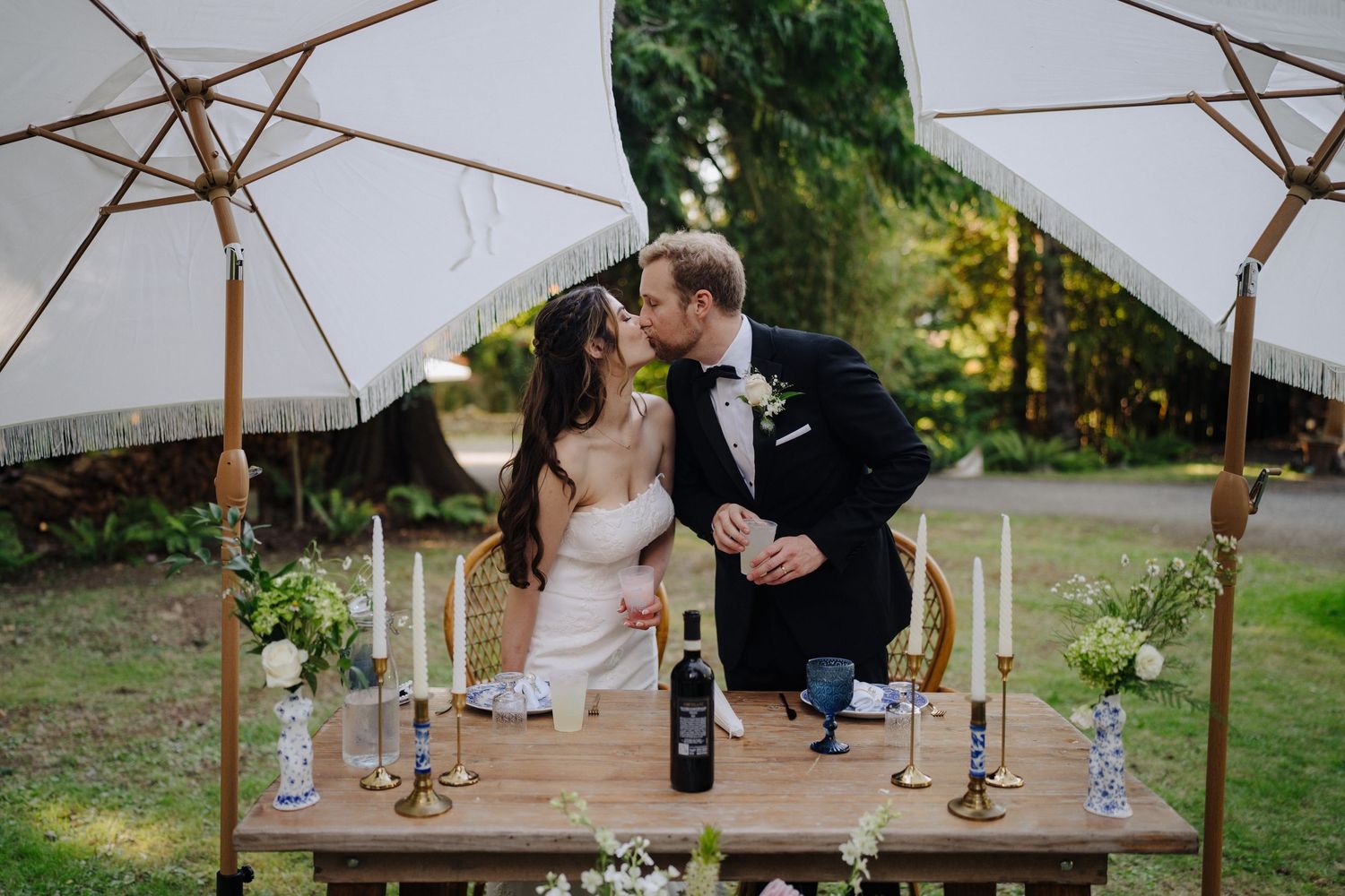 Wedding reception seating area with white umbrellas and wooden tables in garden setting.