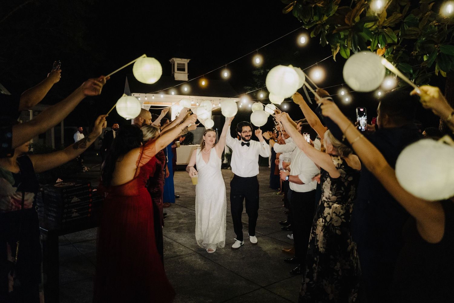 Wedding celebration with guests holding illuminated balloons creating a light tunnel at night.