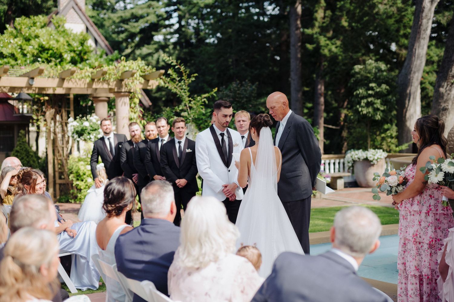 Wedding guests seated outdoors in white chairs watch a ceremony taking place in a lush garden setting.