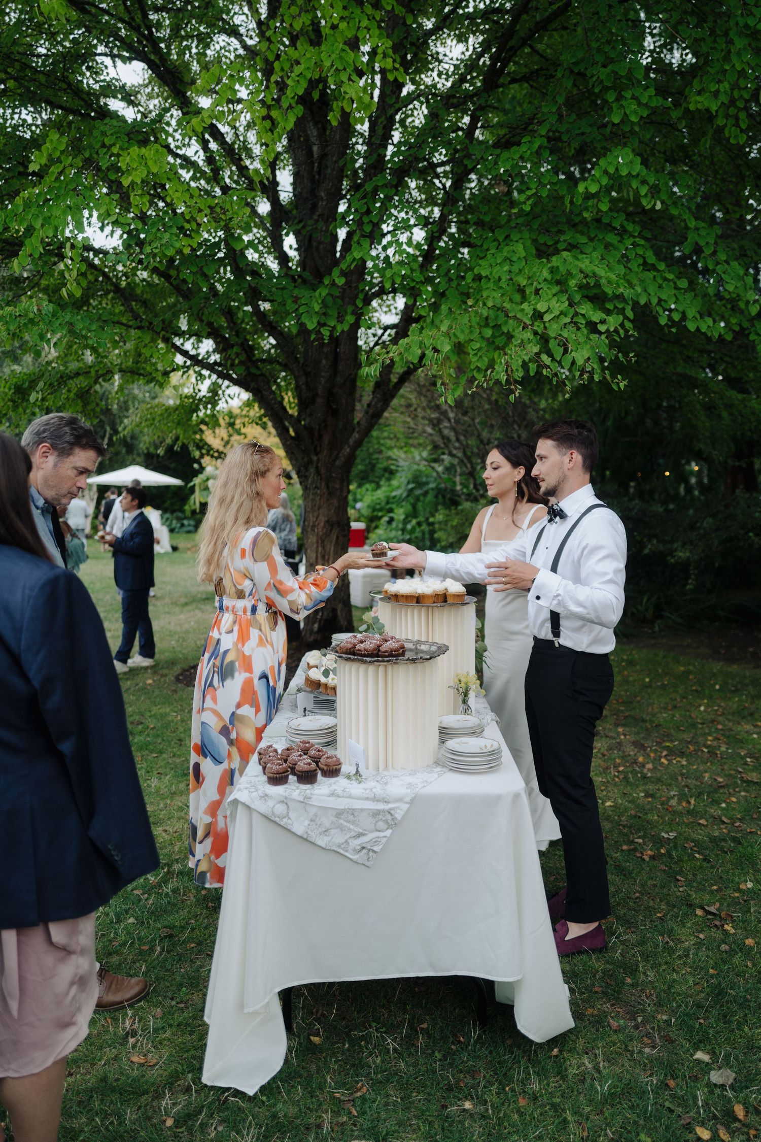 Wedding guests gather around a dessert table set up outdoors under green trees.