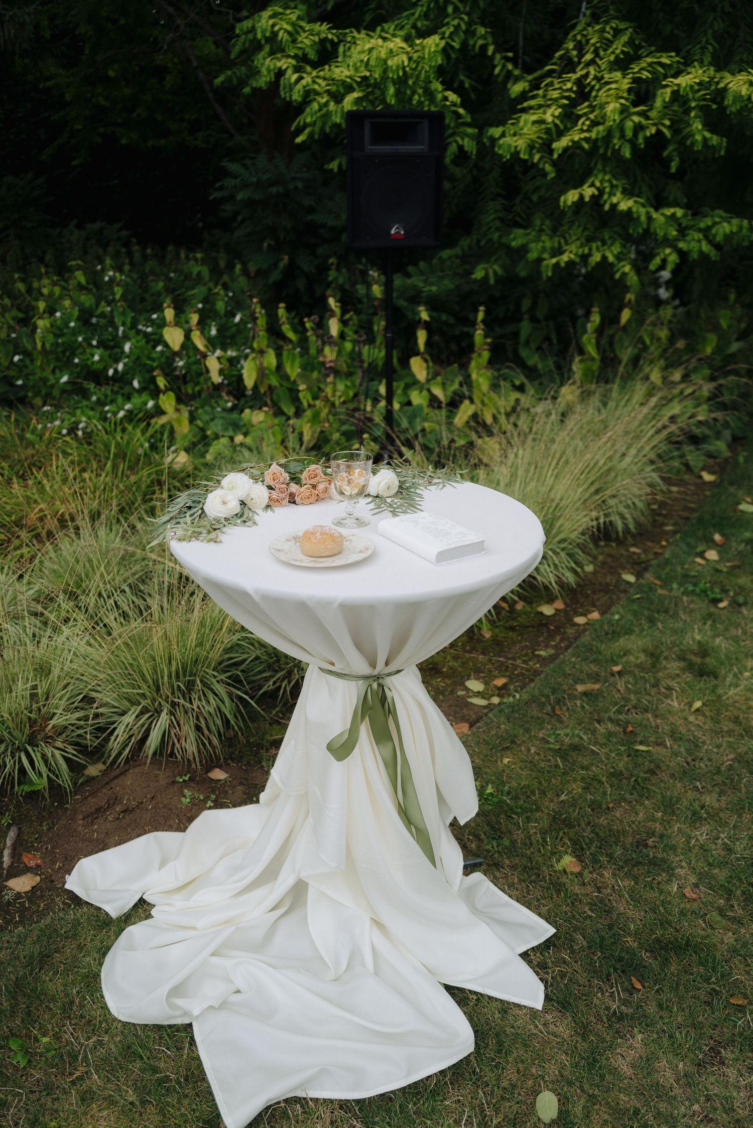 Elegant white cocktail table with green bow sash and decorative settings in an outdoor garden setting.