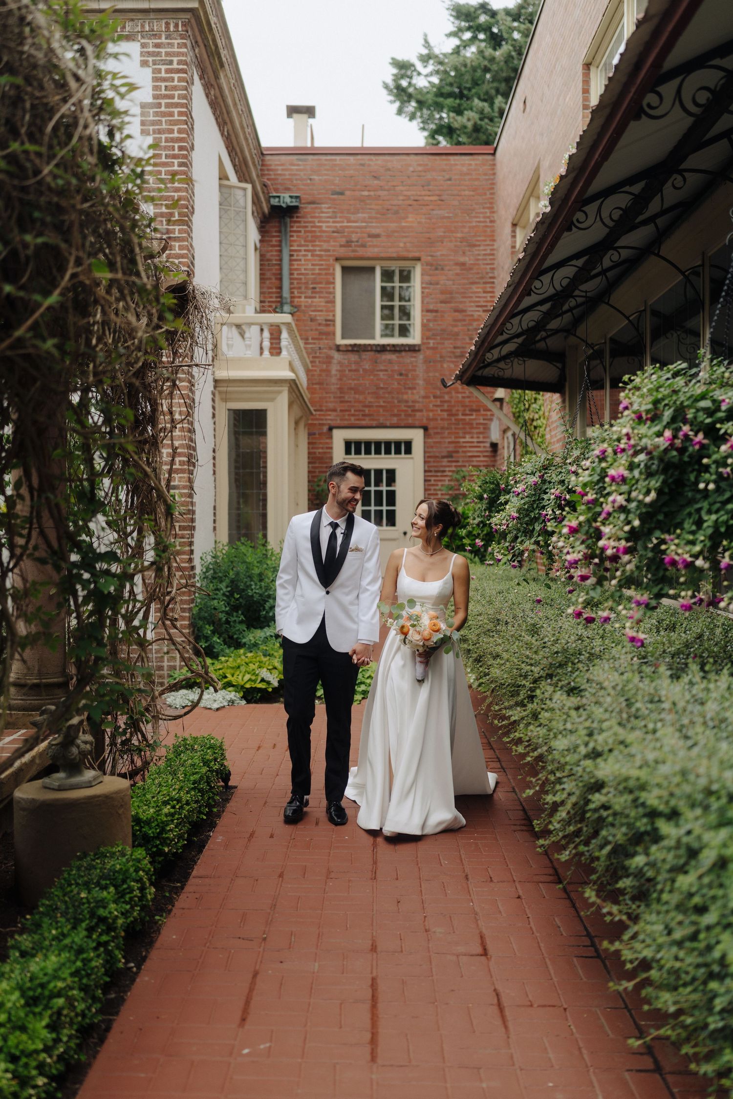 Couple walking down a brick pathway lined with lush greenery and flowers at an elegant venue.