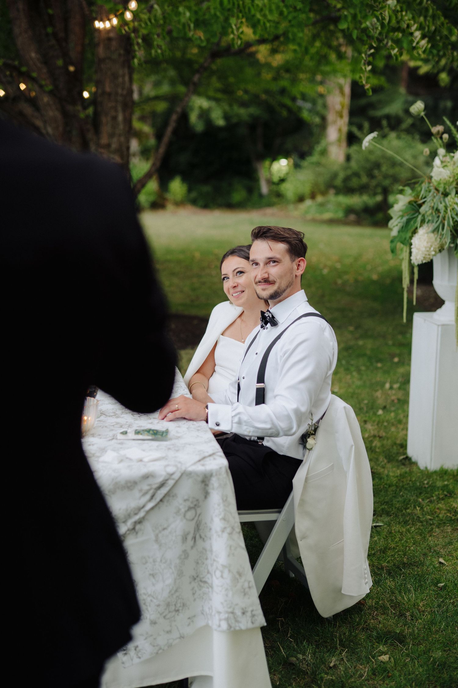A couple shares a joyful moment on white chairs in a garden setting with string lights.