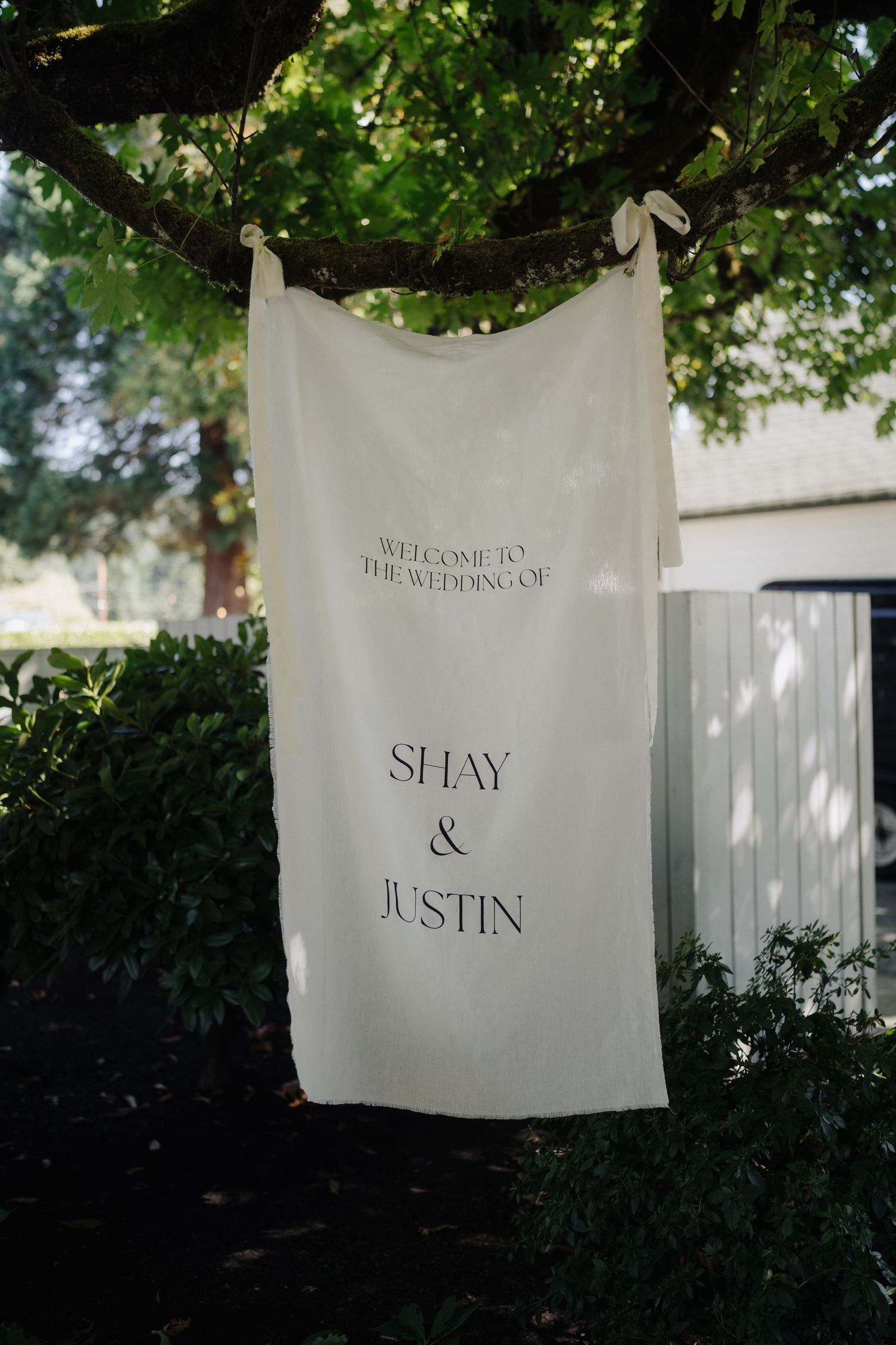 White welcome signs with elegant text hang from tree branches at an outdoor wedding venue.