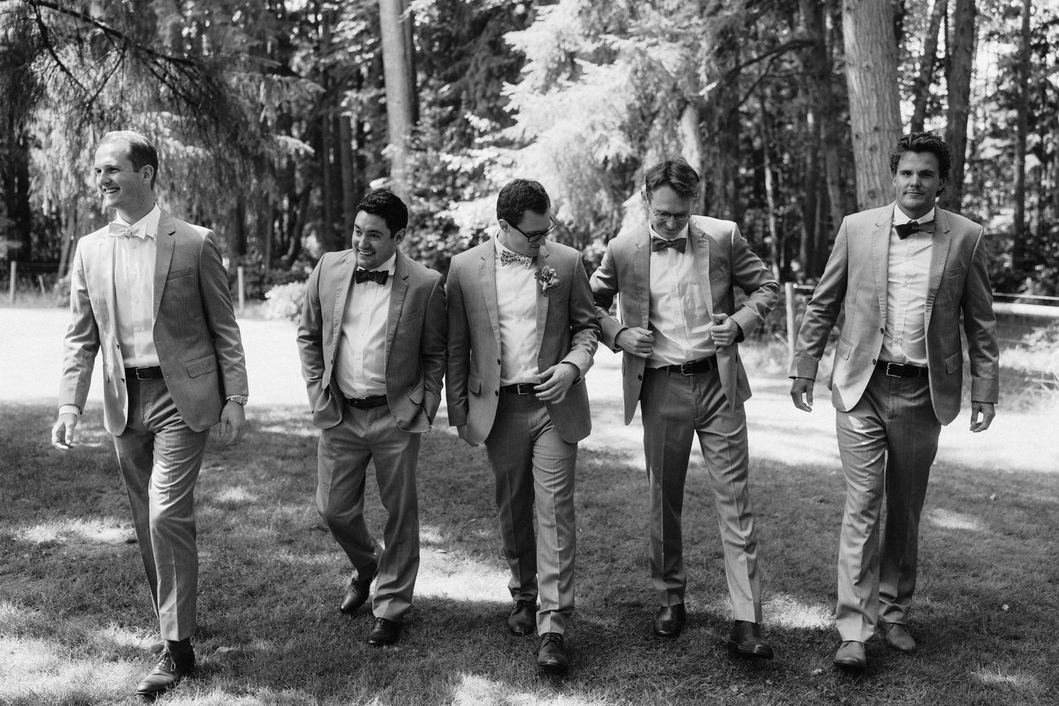 Wedding party walking together through a sunlit forest wearing gray suits and bow ties.