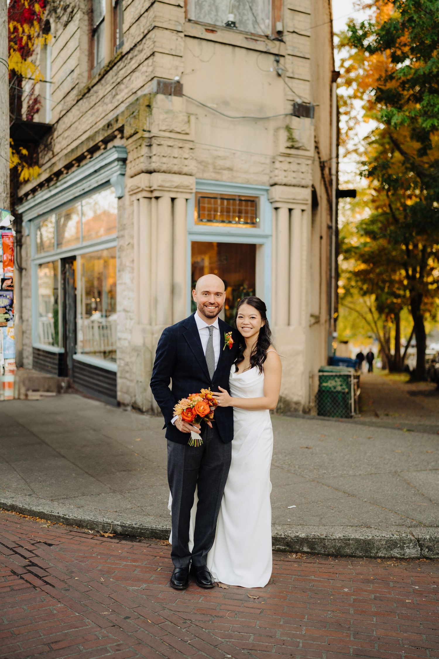 Newlyweds pose outside historic building corner on brick sidewalk with autumn foliage.