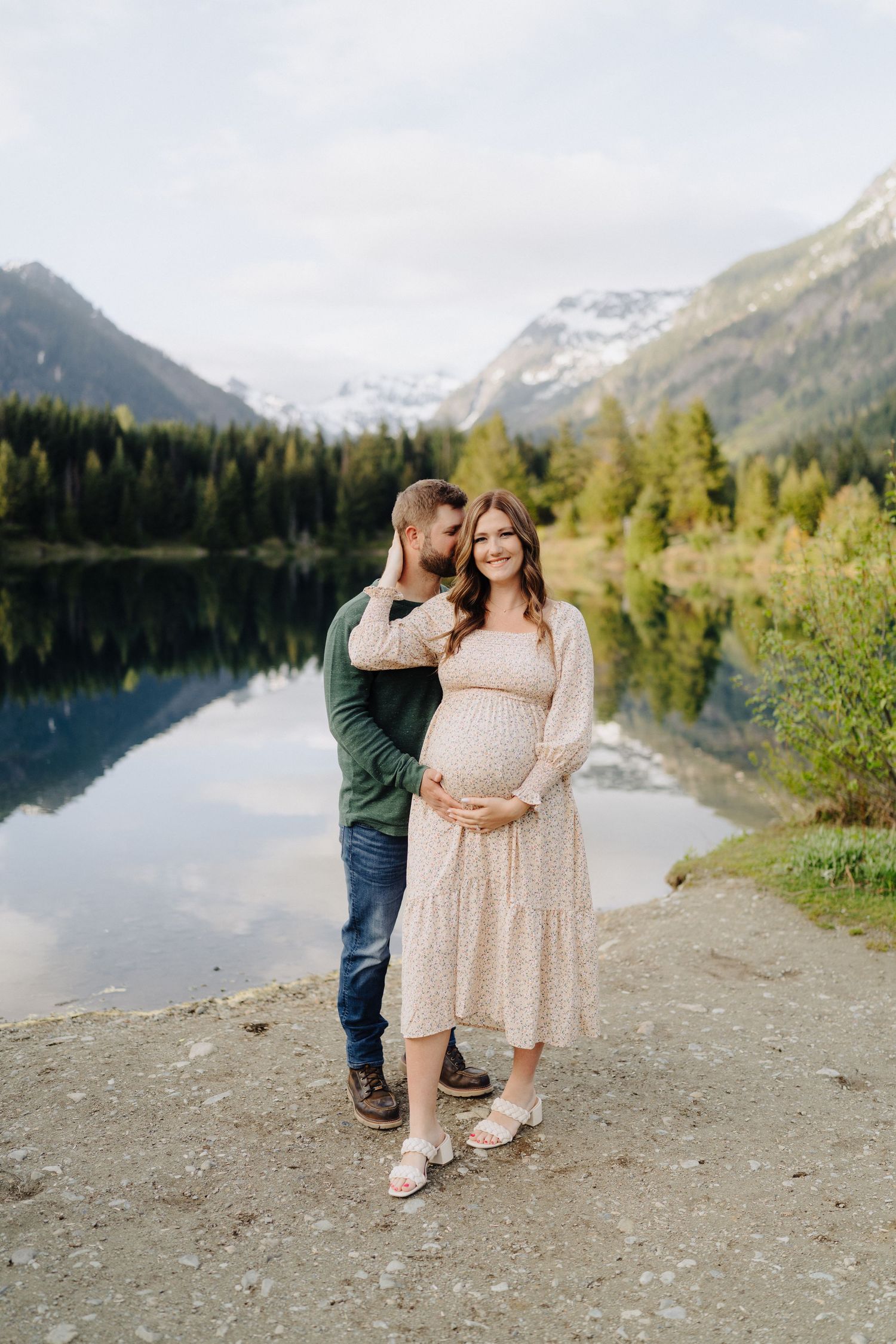 A couple poses together at a scenic mountain lake with snow-capped peaks and evergreen trees in the background.