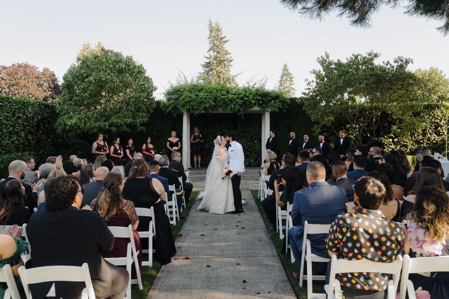 An outdoor wedding ceremony takes place under a white arch with guests seated in white chairs on a stone pathway.