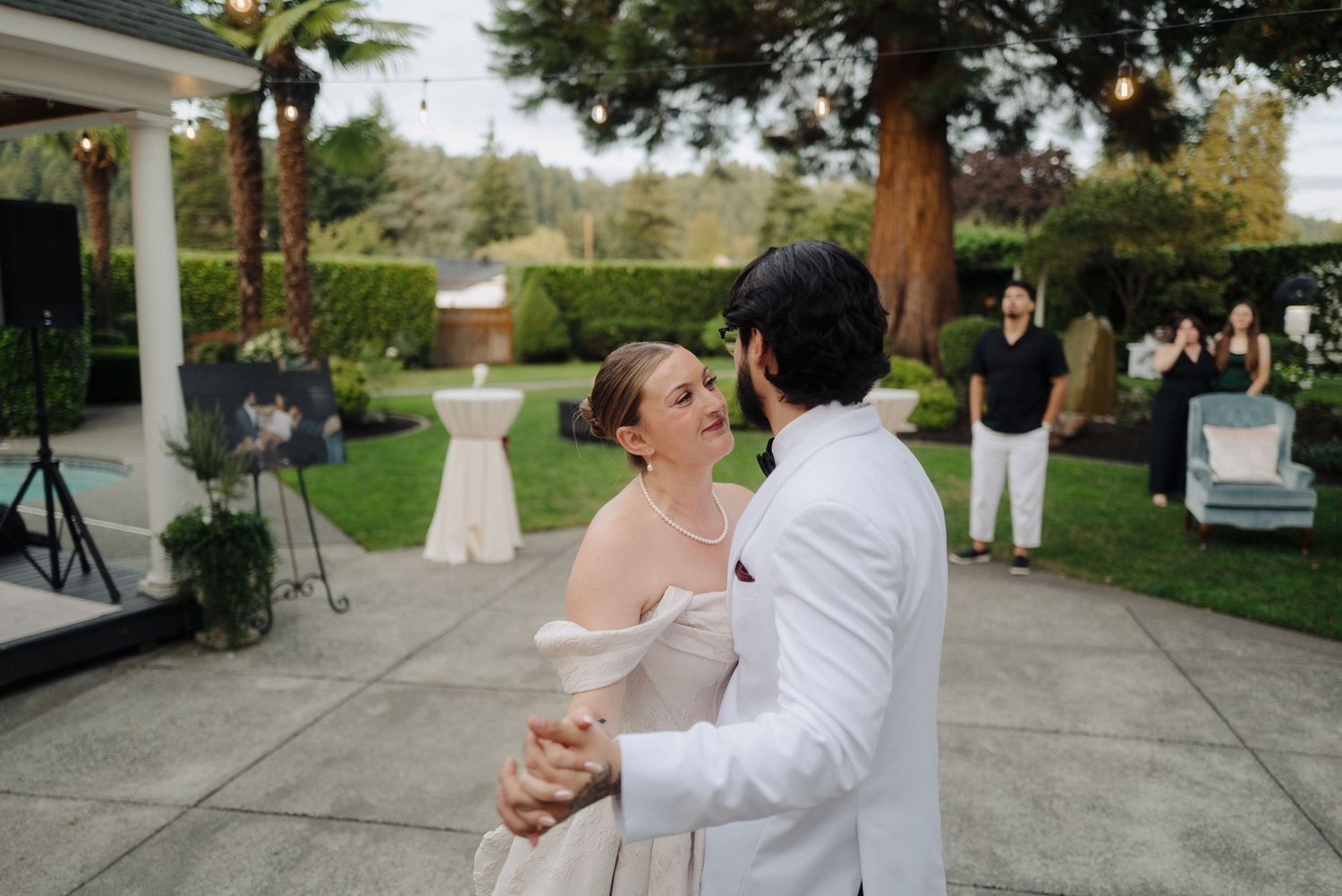 Guests gather for an outdoor wedding reception under tall trees with white tent coverings.