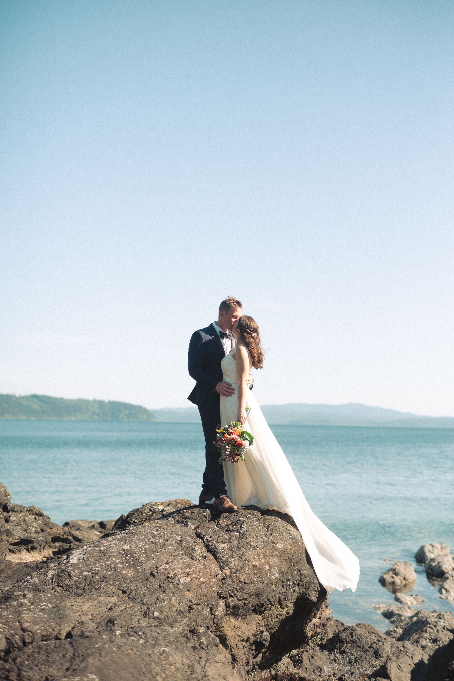 A bride and groom share a romantic kiss on rocky shoreline with blue ocean and mountains in the background.