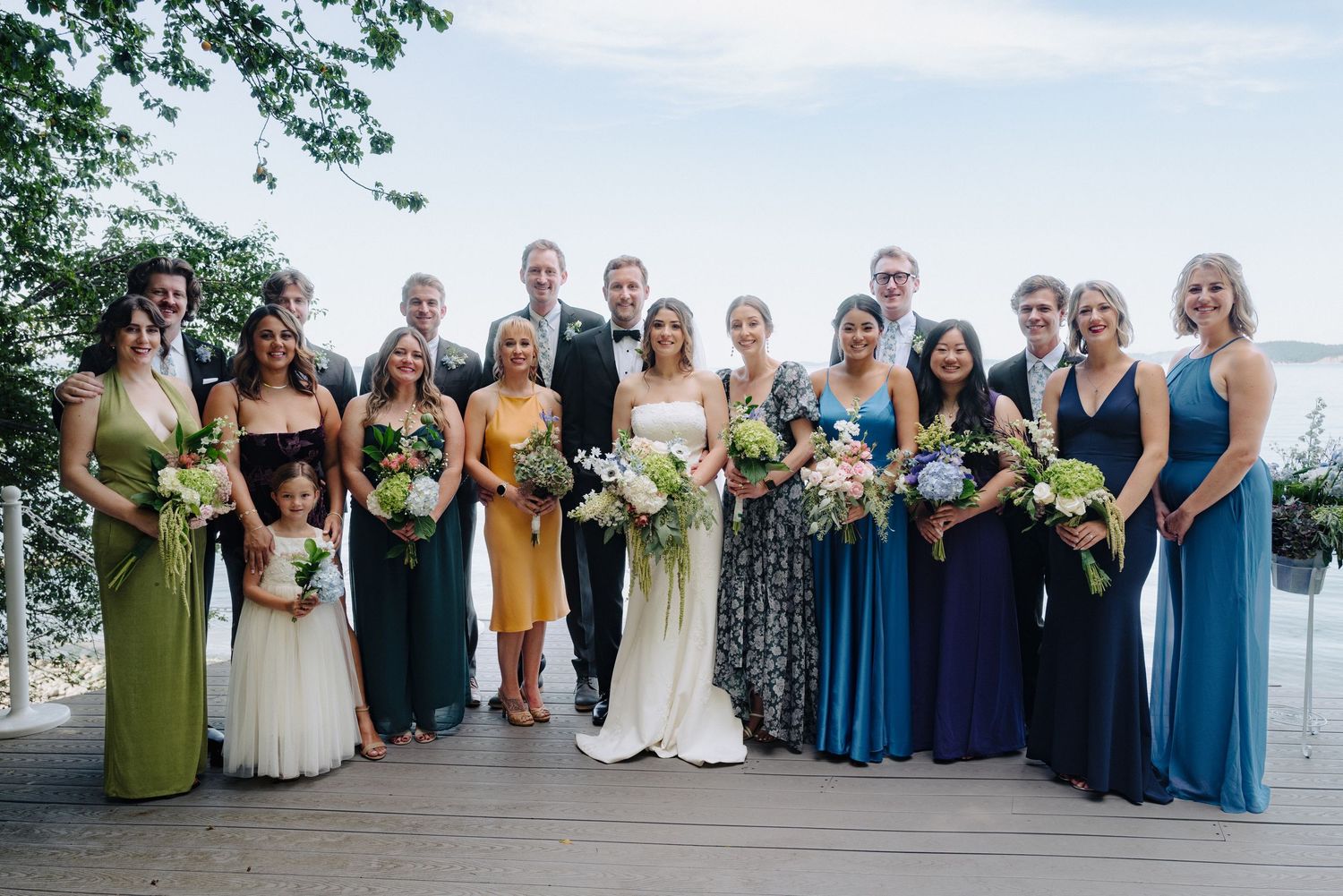 Wedding party members in coordinating blue and green dresses pose on a scenic waterfront deck.