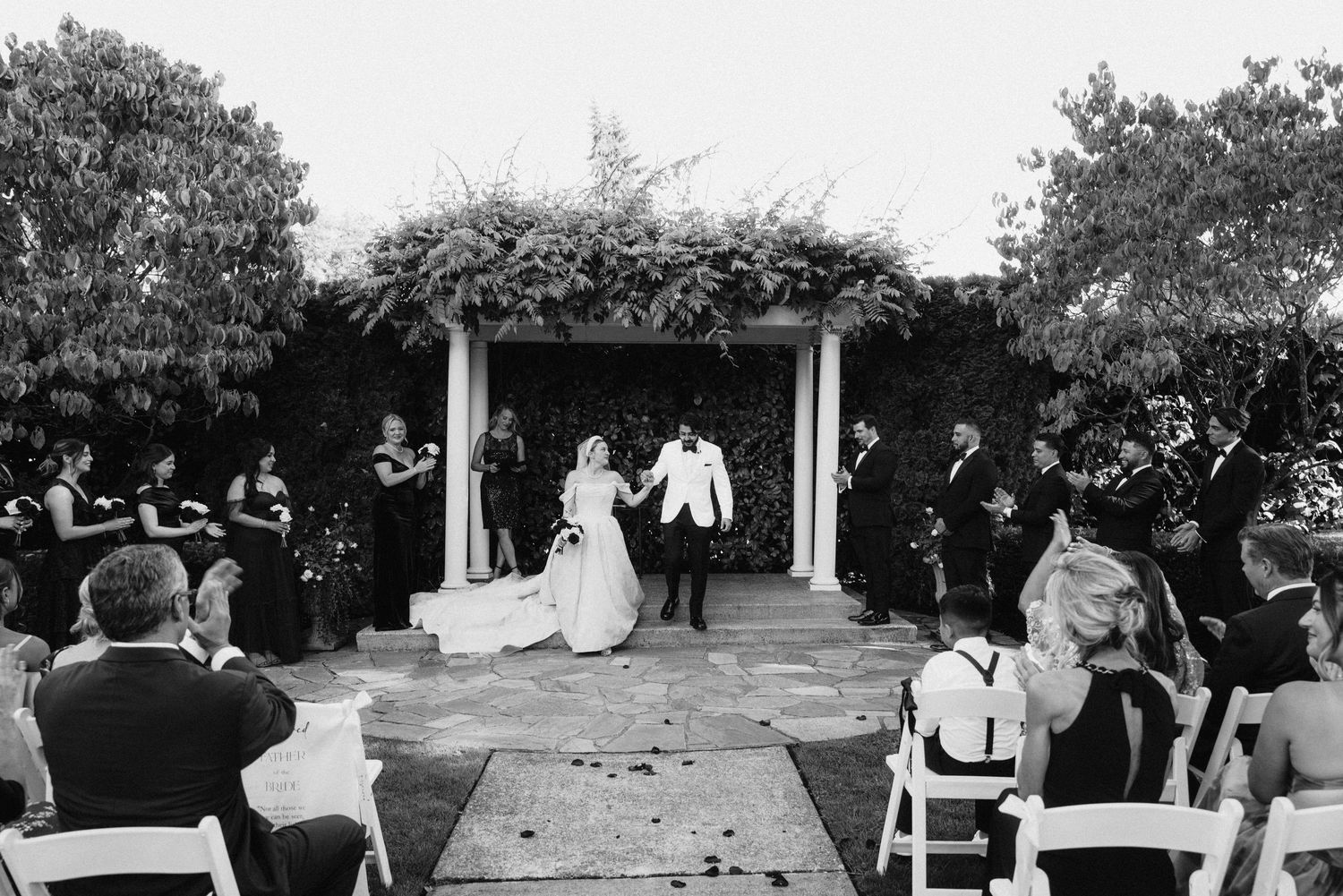 Black and white photo of an outdoor wedding ceremony with white chairs and floral arrangements.