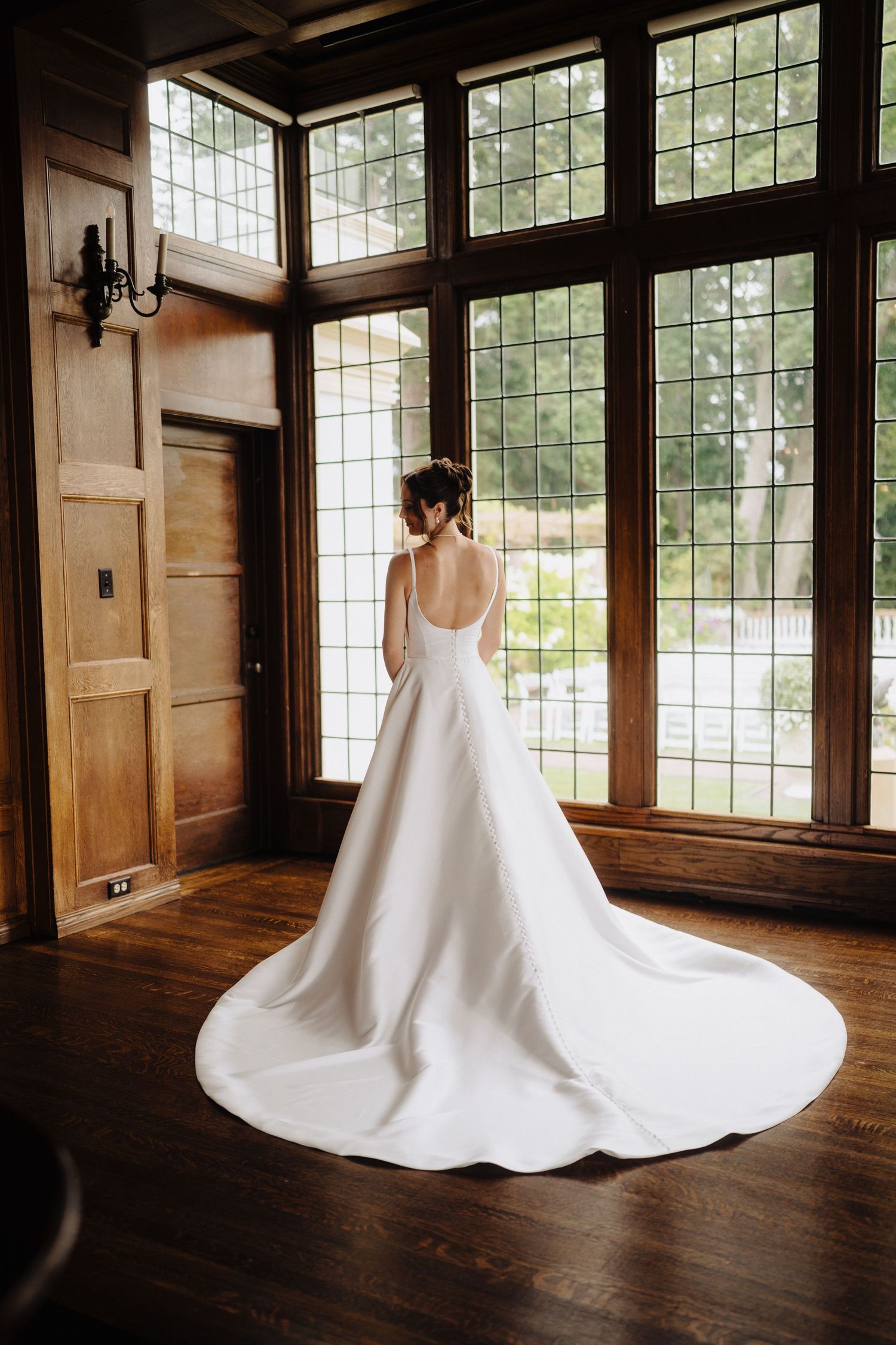 Elegant white wedding dress with long train photographed from behind in a sunlit room with wooden floors and large windows.