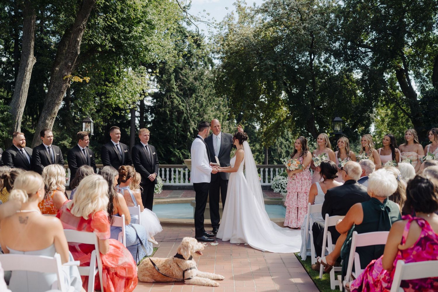 Wedding ceremony in progress under shady trees with guests seated in white chairs.