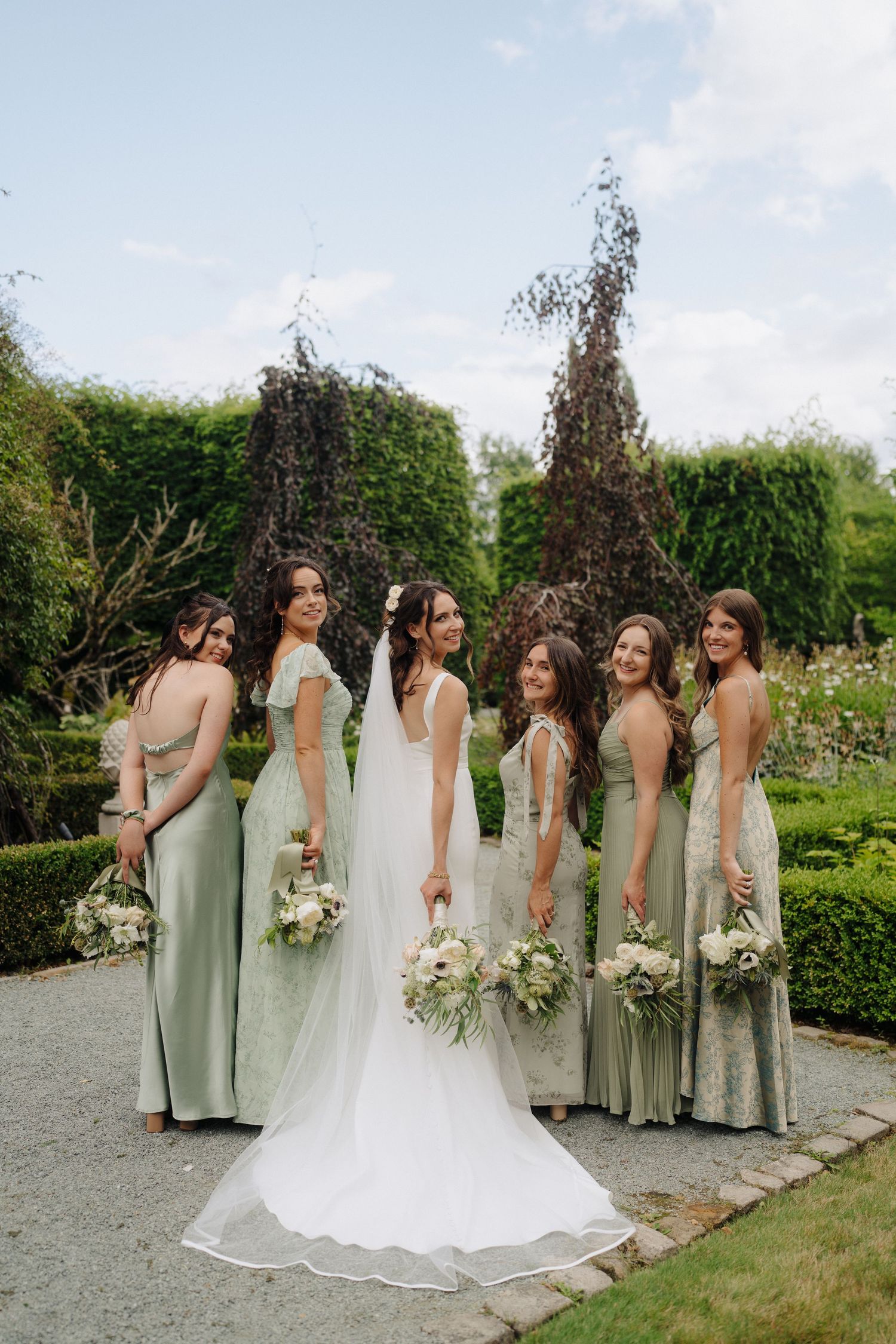 Bridesmaids in sage green dresses posing with bride in white gown in a garden setting.