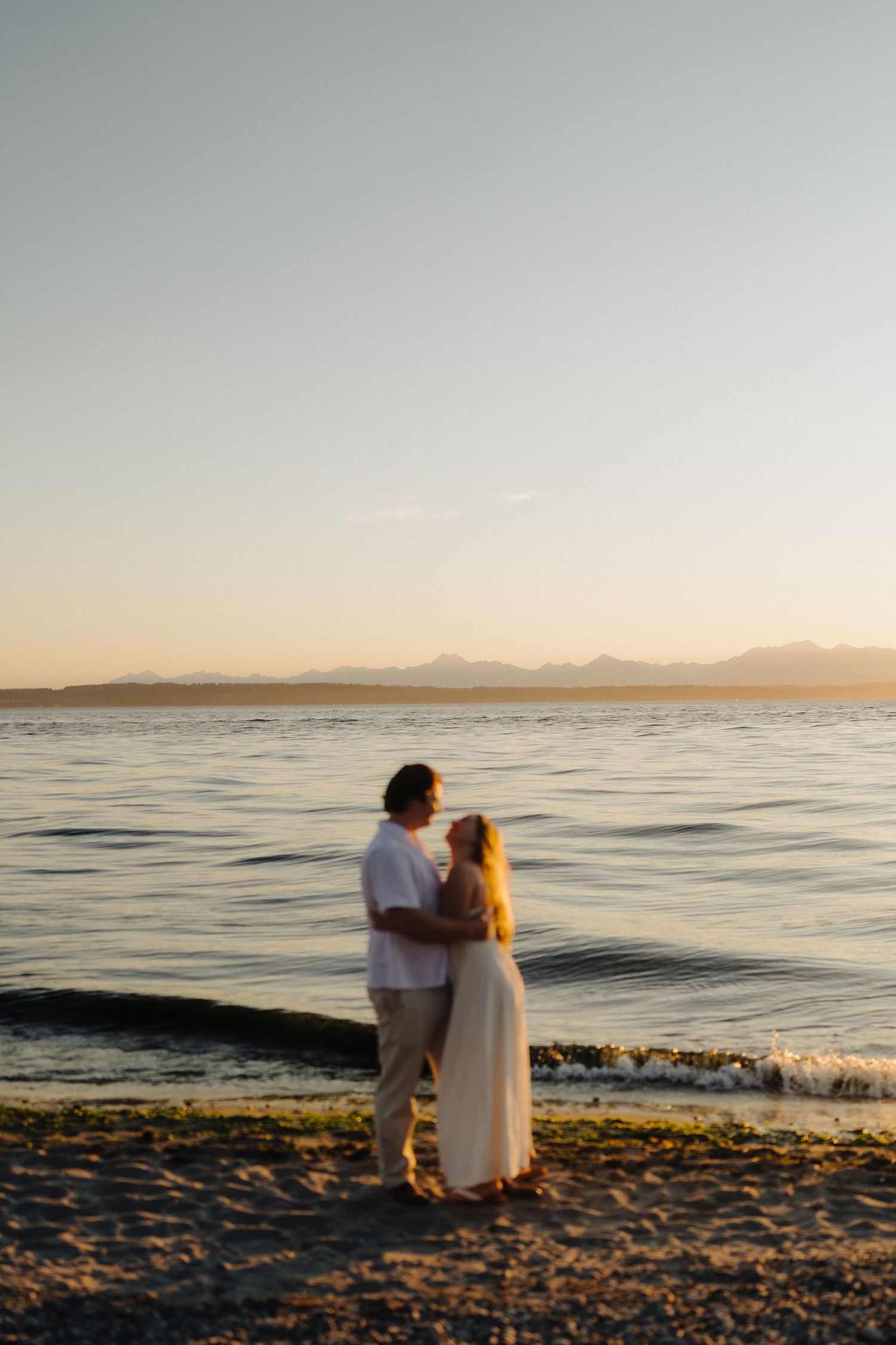 A couple in white clothing sits together on a beach at sunset with mountains visible across the water.