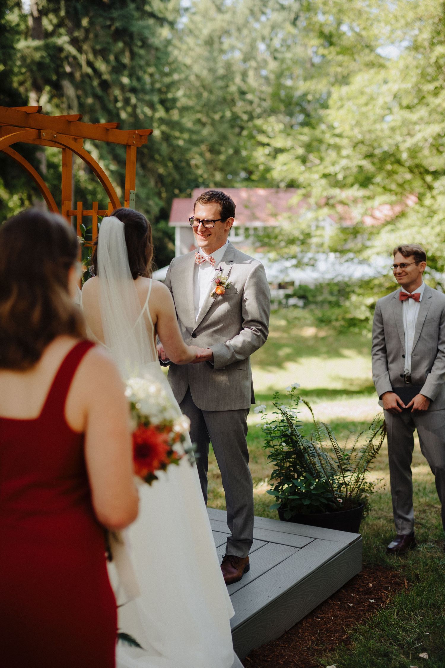 Wedding couple exchanges vows on wooden platform with rustic wooden arch in wooded outdoor setting.