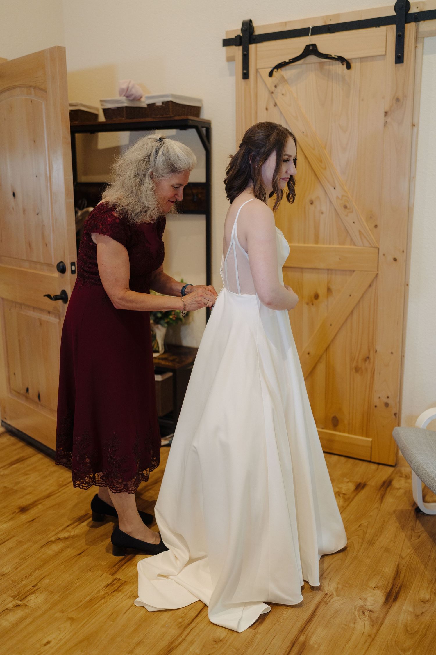 A wedding dress fitting shows final adjustments being made to an elegant white gown in a rustic dressing room.