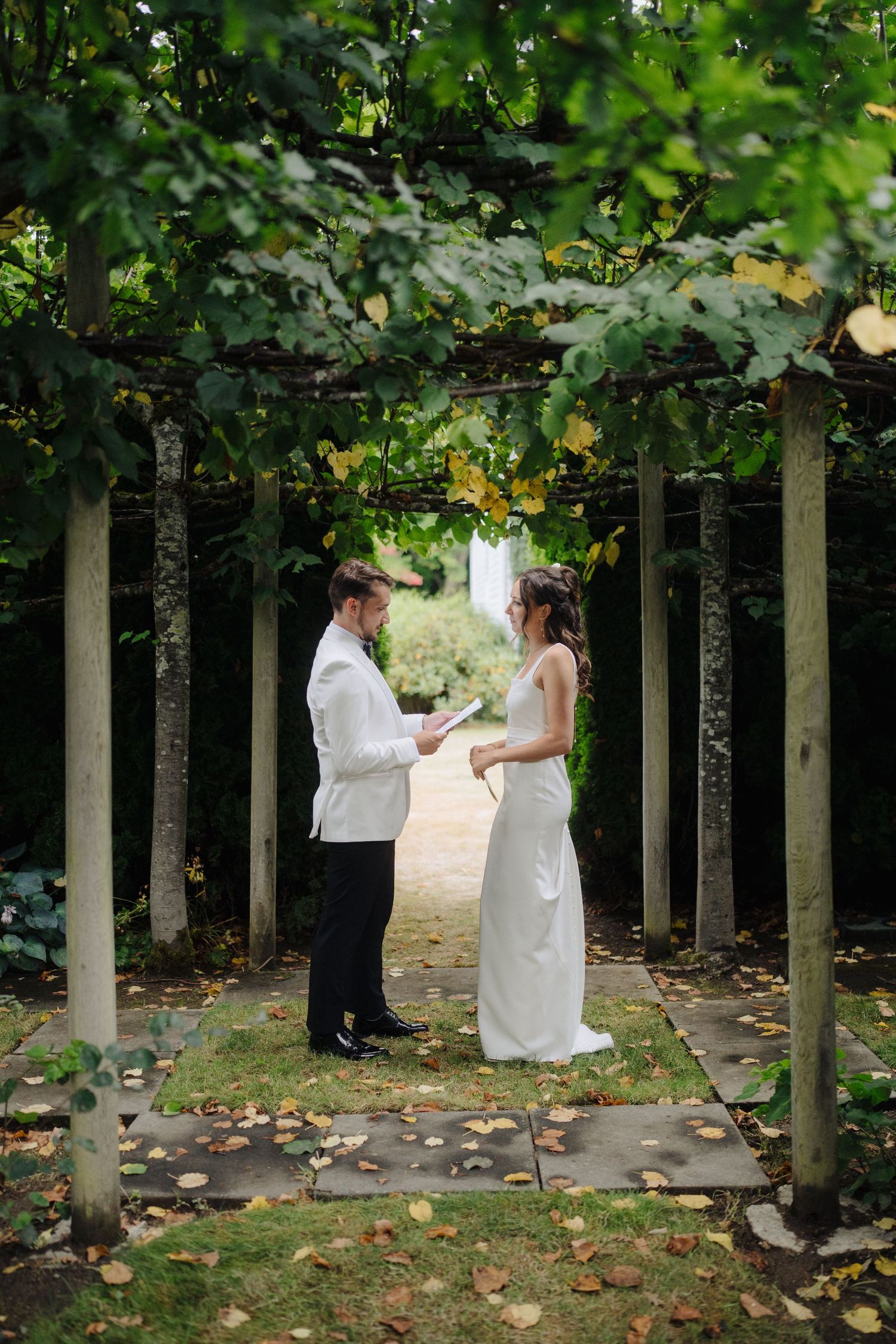 Wedding couple walks together through a scenic garden path lined with tall trees.